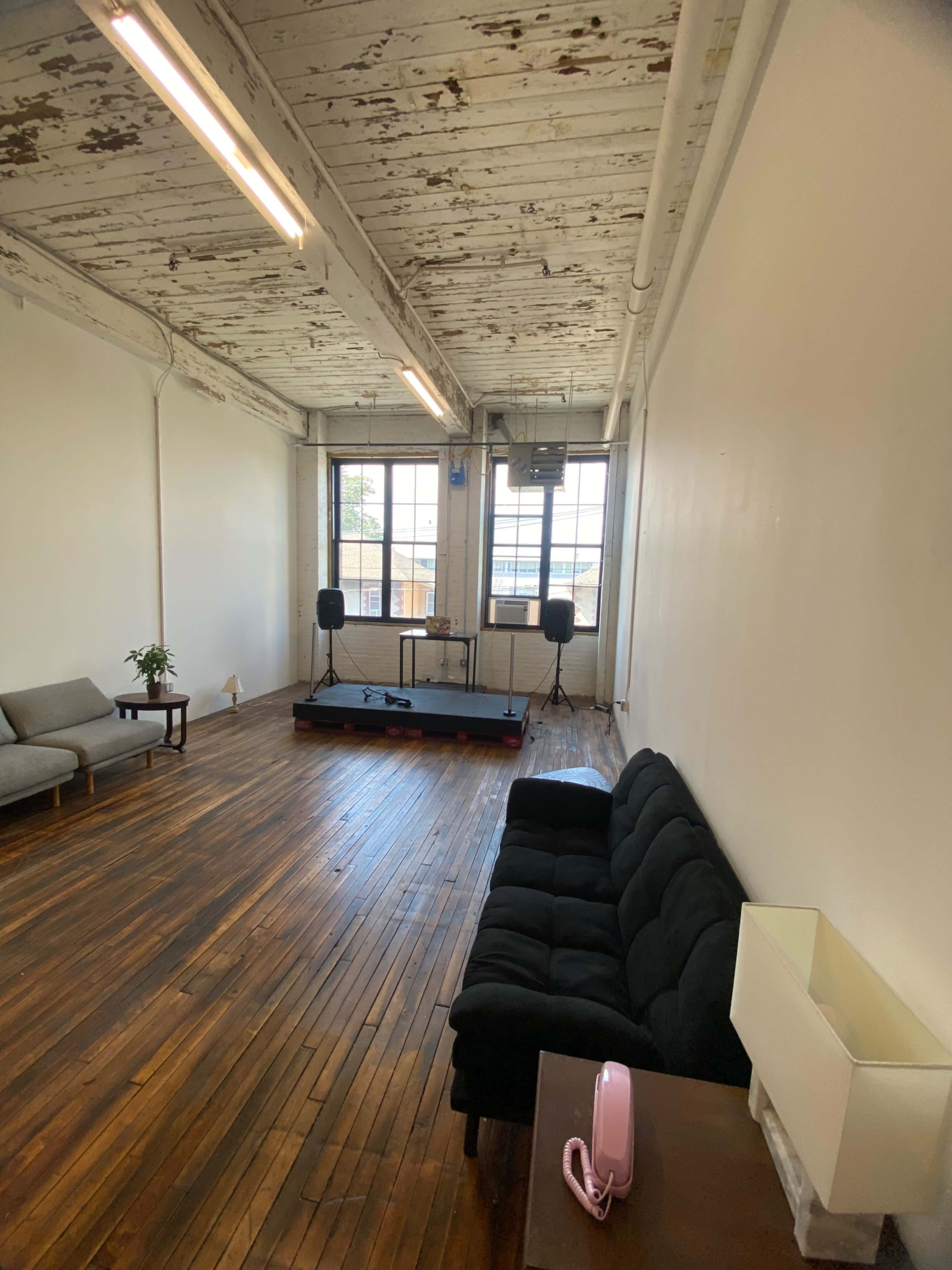 The image shows an empty room with wooden floors, a sofa on one side, and a small stage with microphones set up in front of a large window.