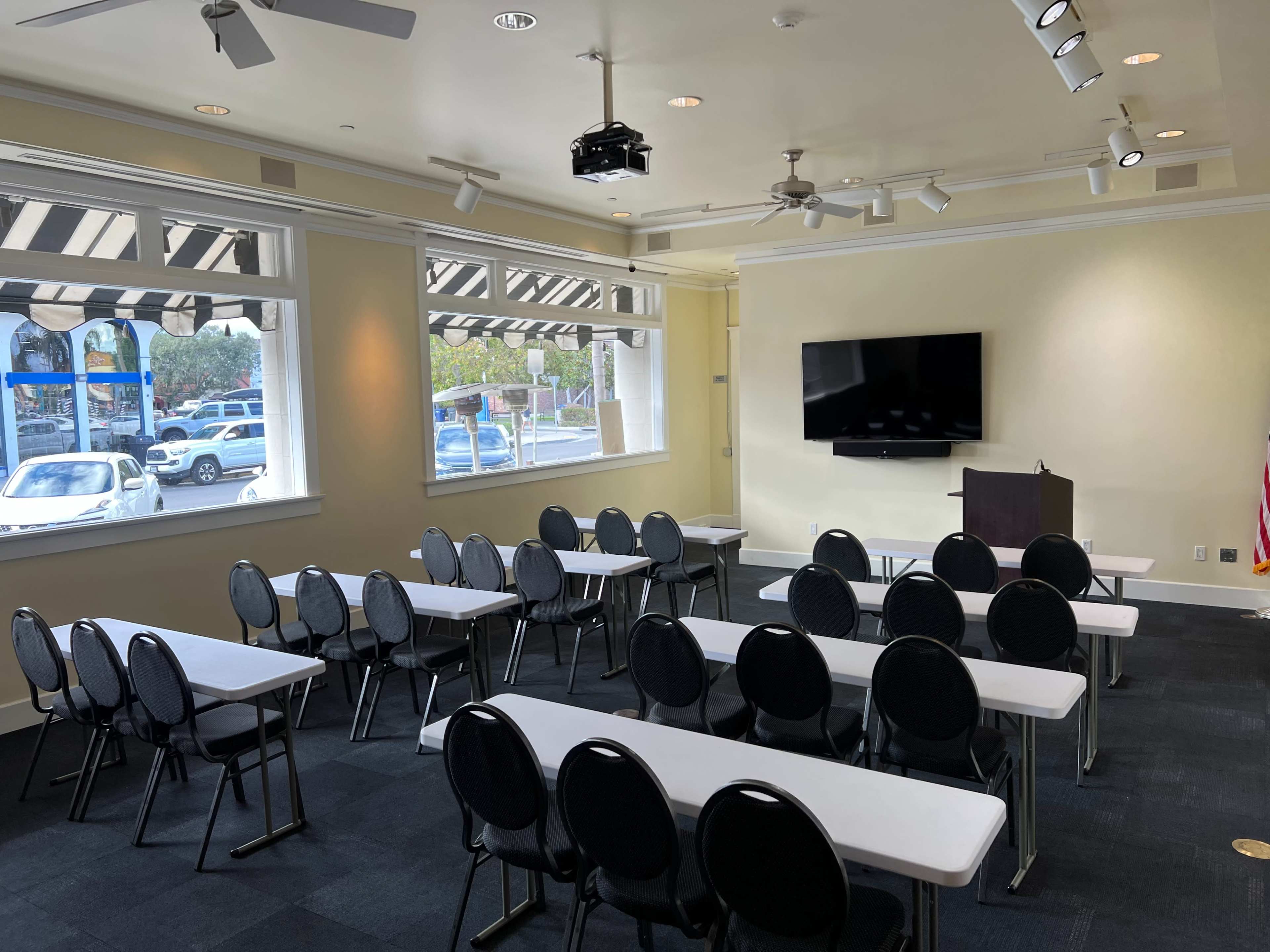 A room set up for a presentation with several rows of tables and chairs, a podium, and a large screen on one wall.