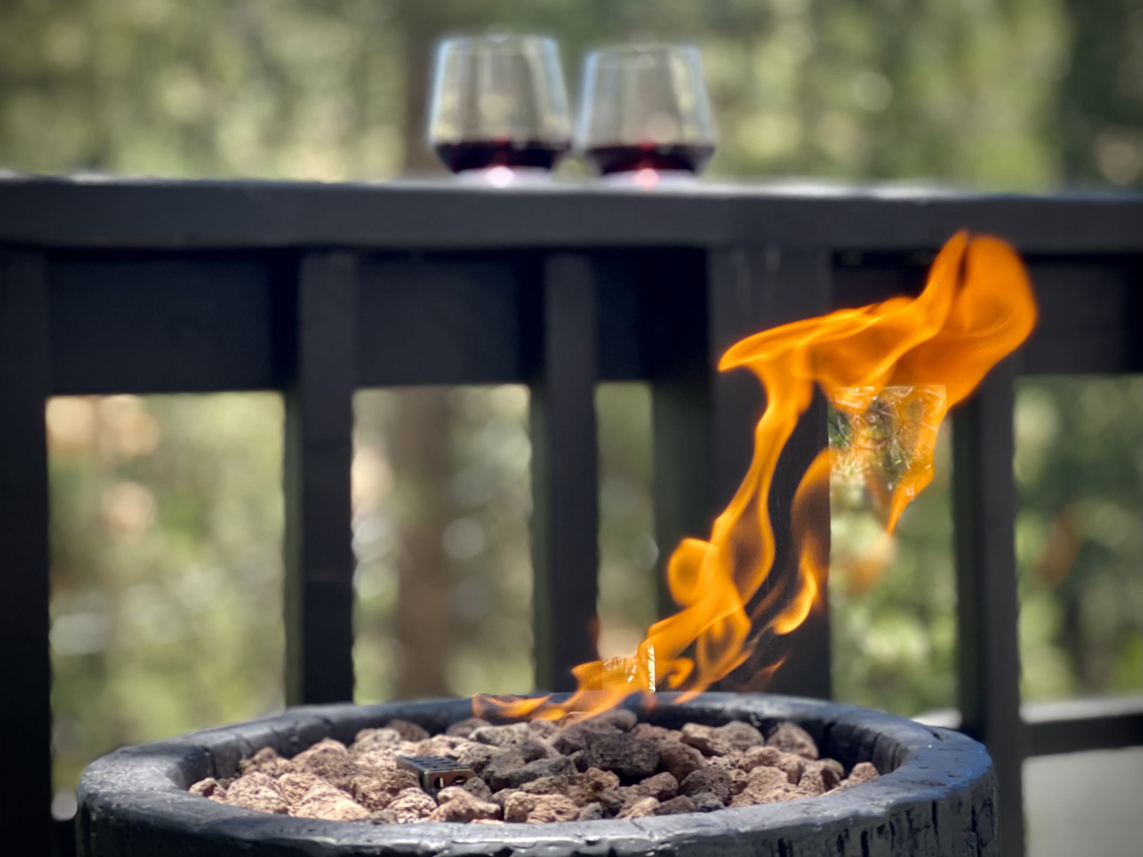 A lit fire pit with flames rising above a bed of rocks is positioned on a deck, with two glasses of red wine in the background.
