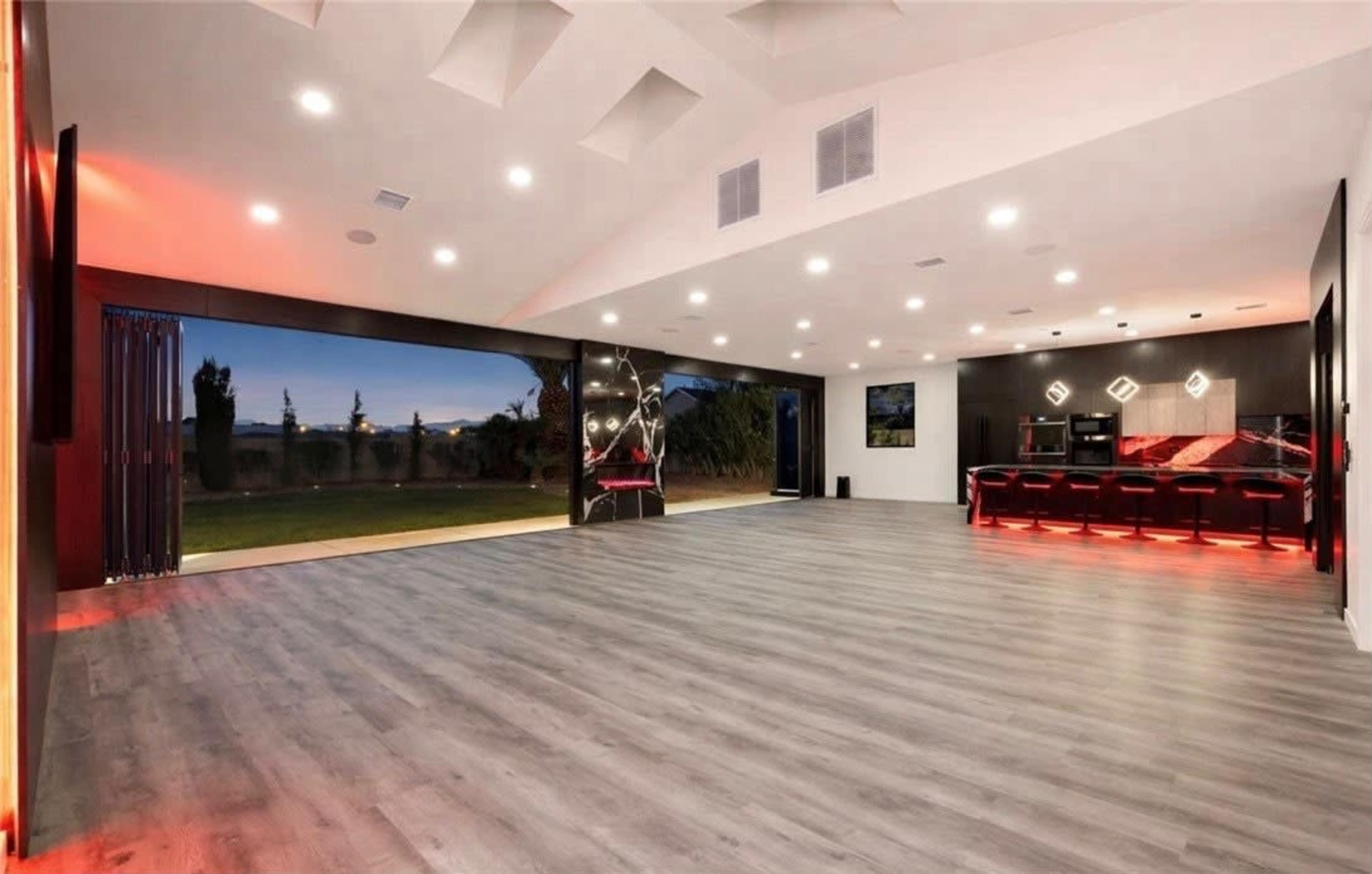 The image shows a spacious, modern interior with grey wooden flooring and large windows offering a view of the outdoors.