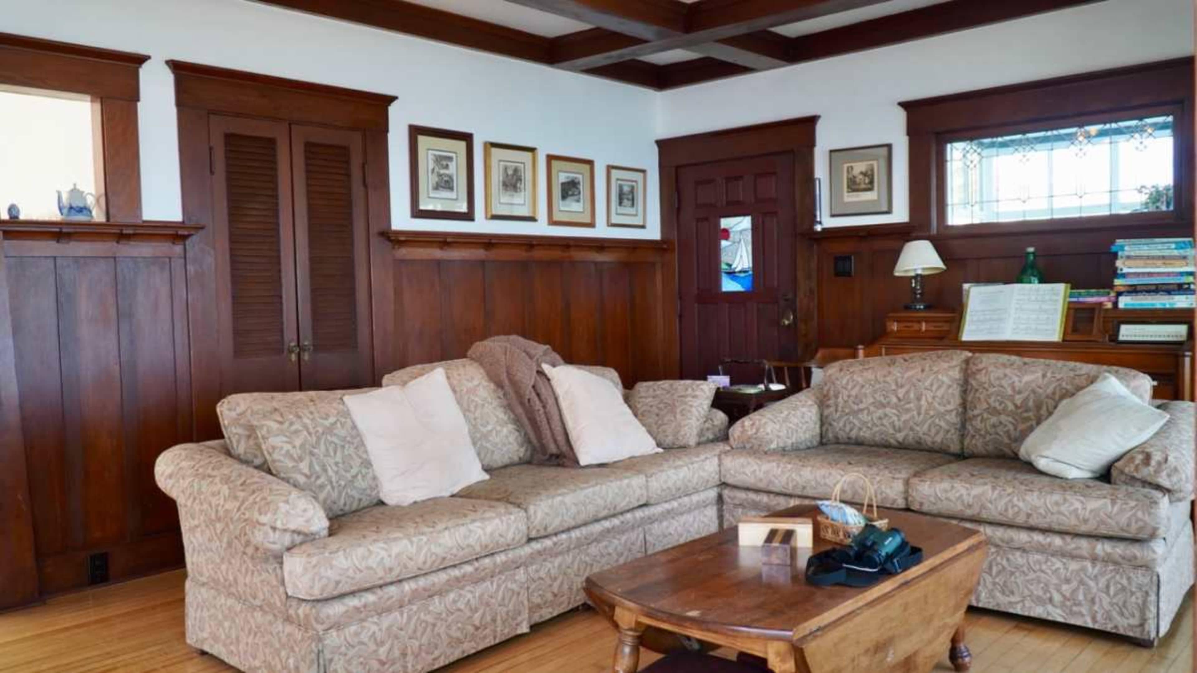 The image shows a living room with a patterned sectional sofa, wooden walls, and a piano against the wall.