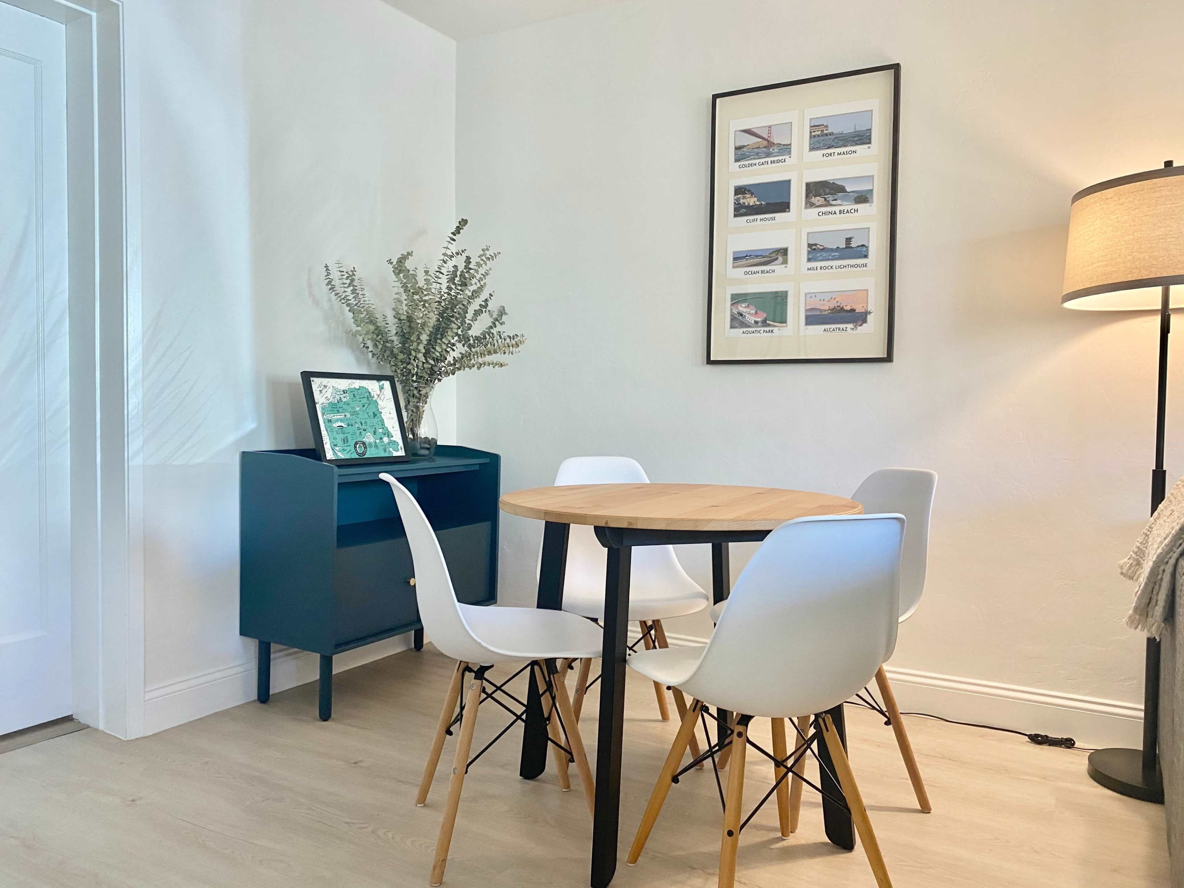 A small dining area features a round wooden table with four white chairs, a blue cabinet, and framed artwork on the wall.