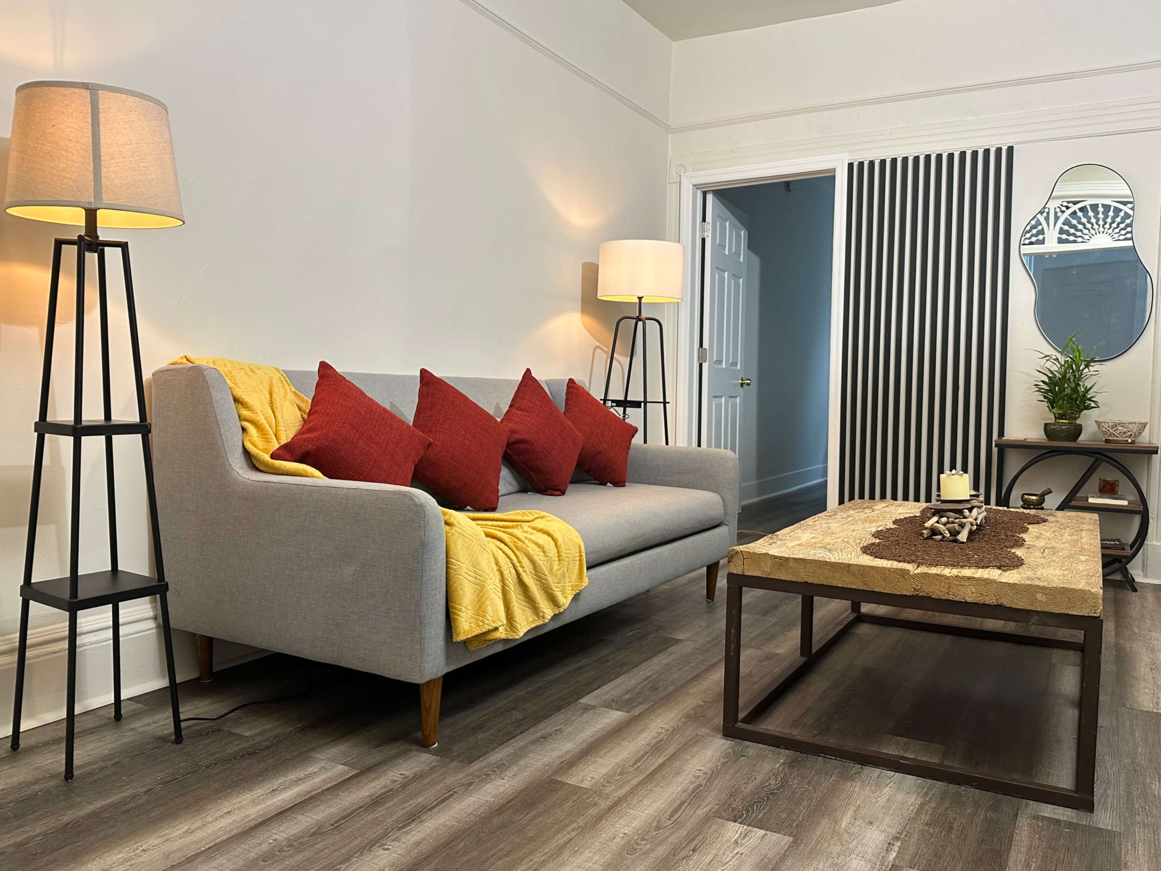 The image shows a living room with a gray sofa adorned with red pillows and a yellow throw blanket, flanked by lamp stands, and featuring a wooden coffee table and a plant in the corner.