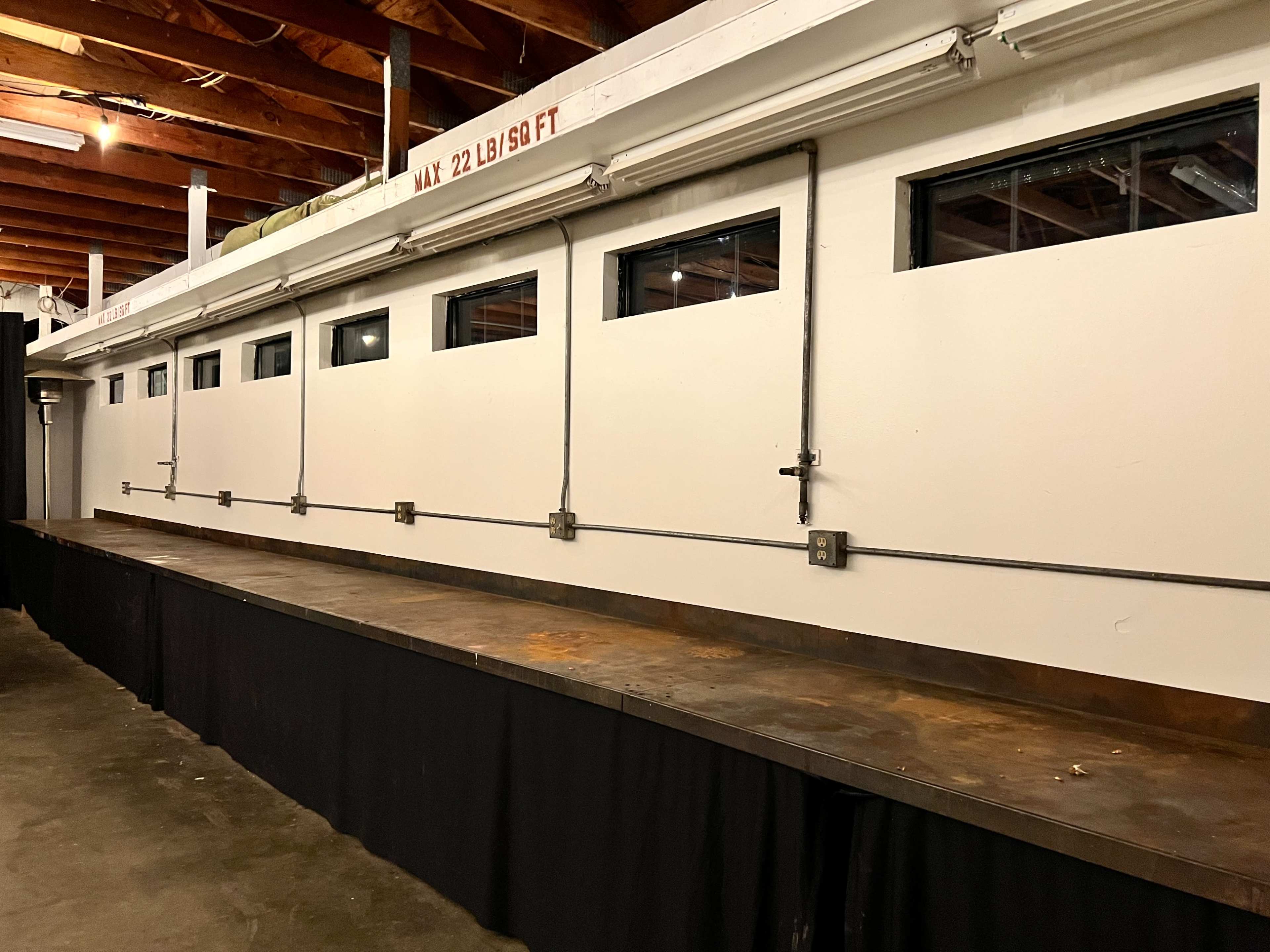 The image shows a long, metal countertop with exposed piping and several small windows above it, located in a room with a ceiling of wooden beams.