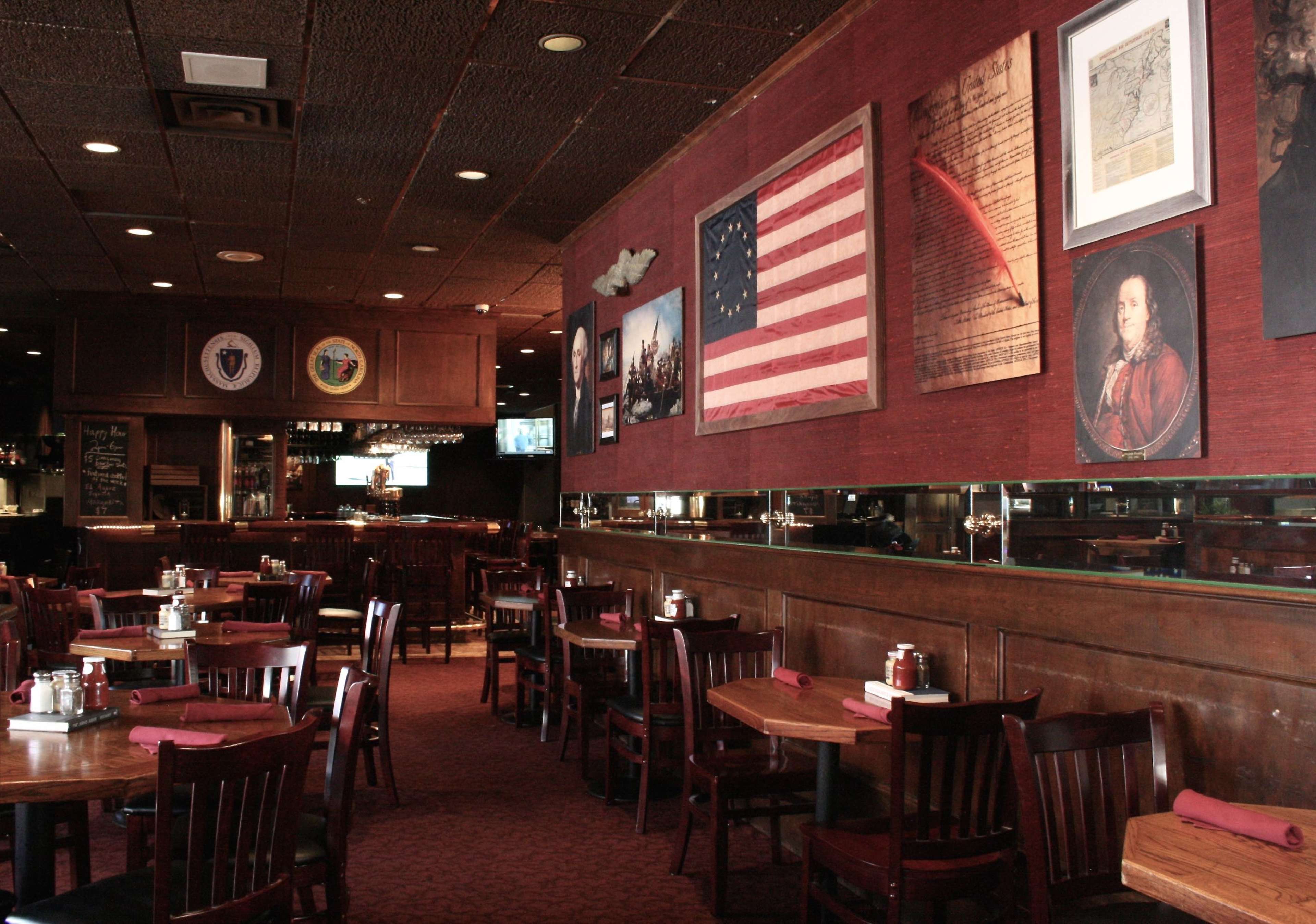A restaurant interior featuring wooden tables and chairs, decorated walls with framed artworks, and visible bar area in the background.