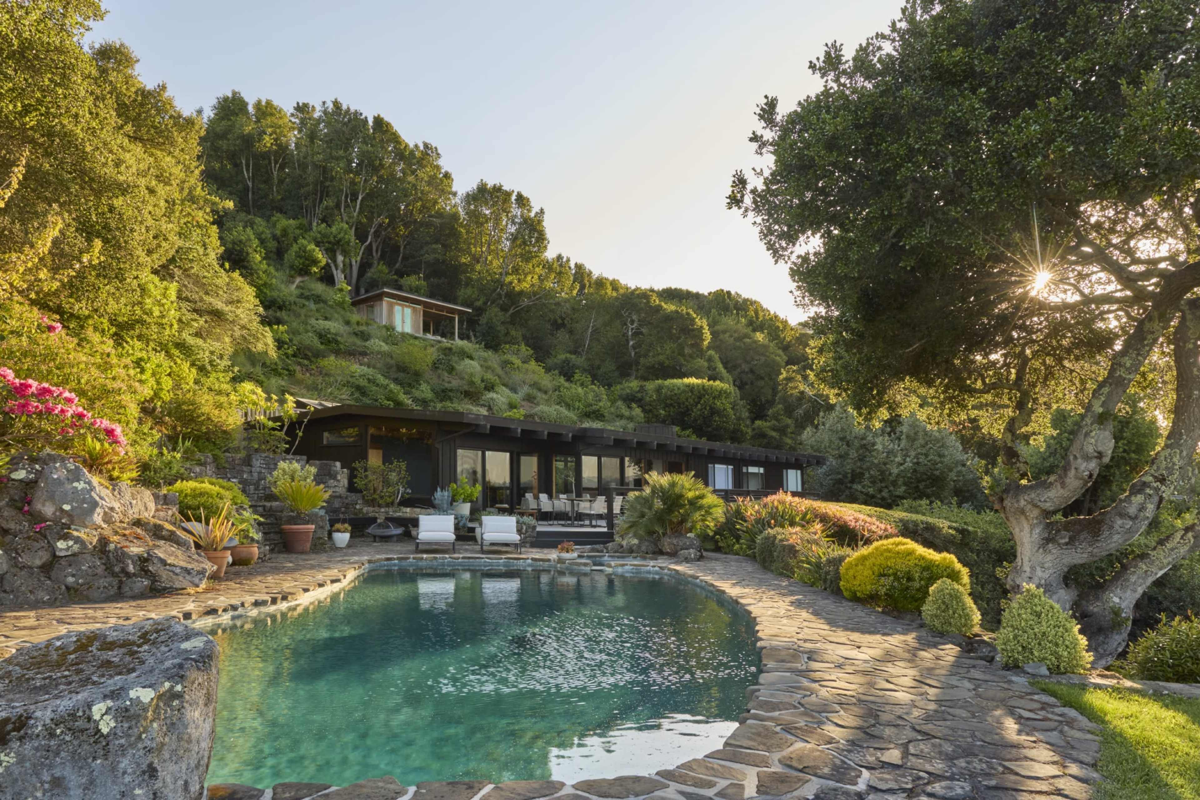 A modern house with large windows overlooks a curved stone pool surrounded by lush greenery and colorful flowers on a hillside.
