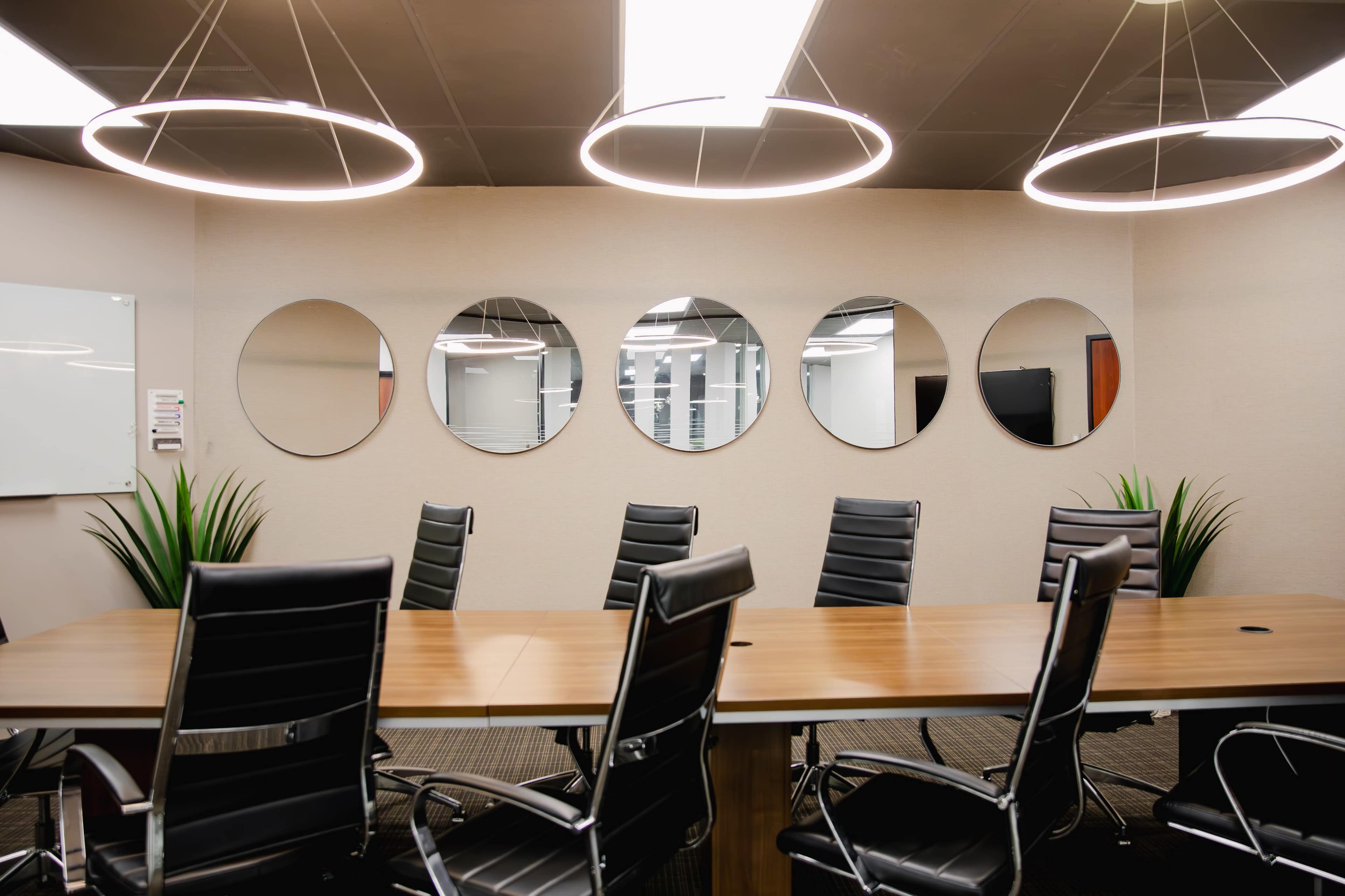 A modern conference room features a long wooden table surrounded by black leather chairs, with five round mirrors mounted on the wall and illuminated by circular pendant lights.