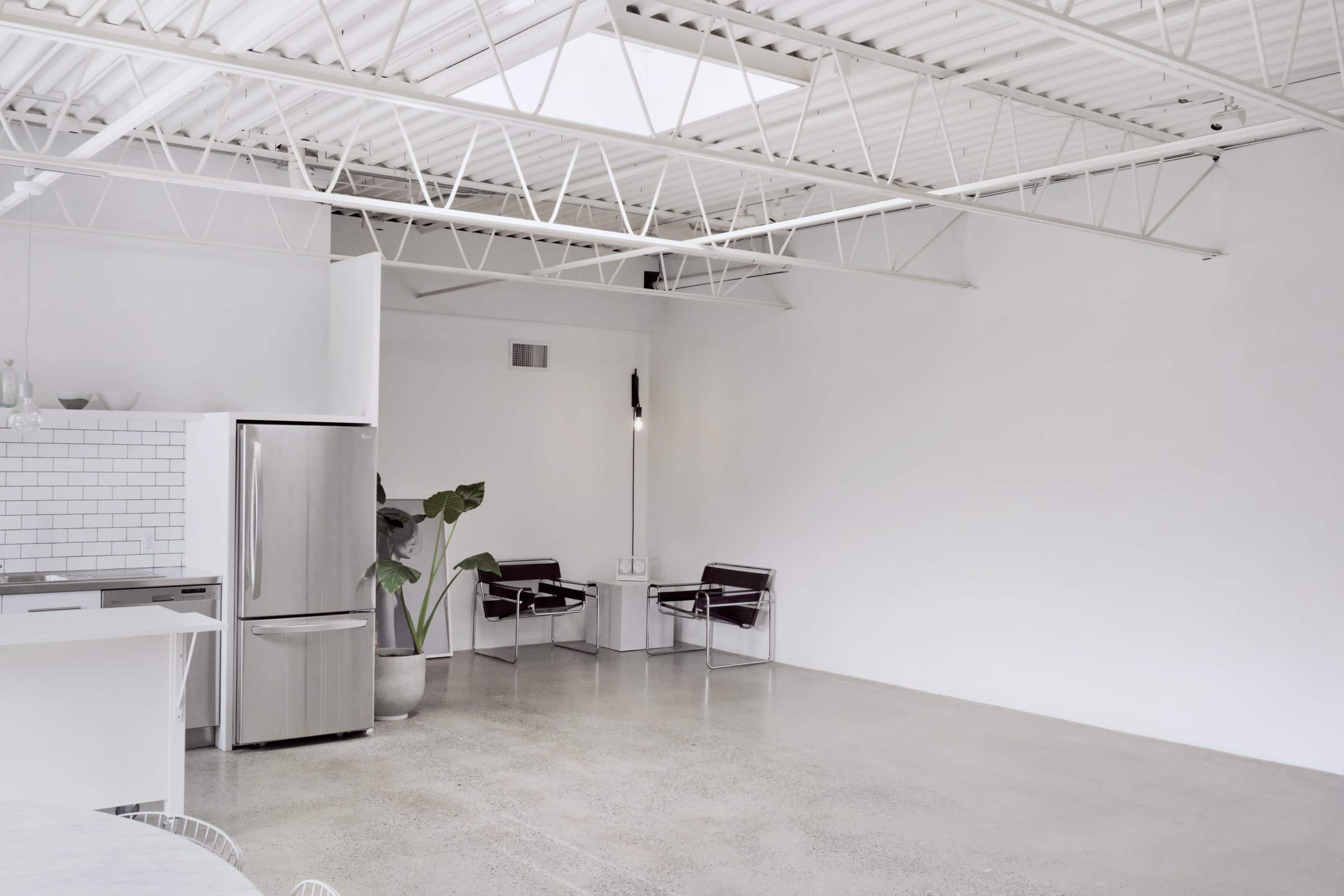 The image shows a spacious, minimalist interior with a stainless steel refrigerator, a small potted plant, and two chairs on a concrete floor.