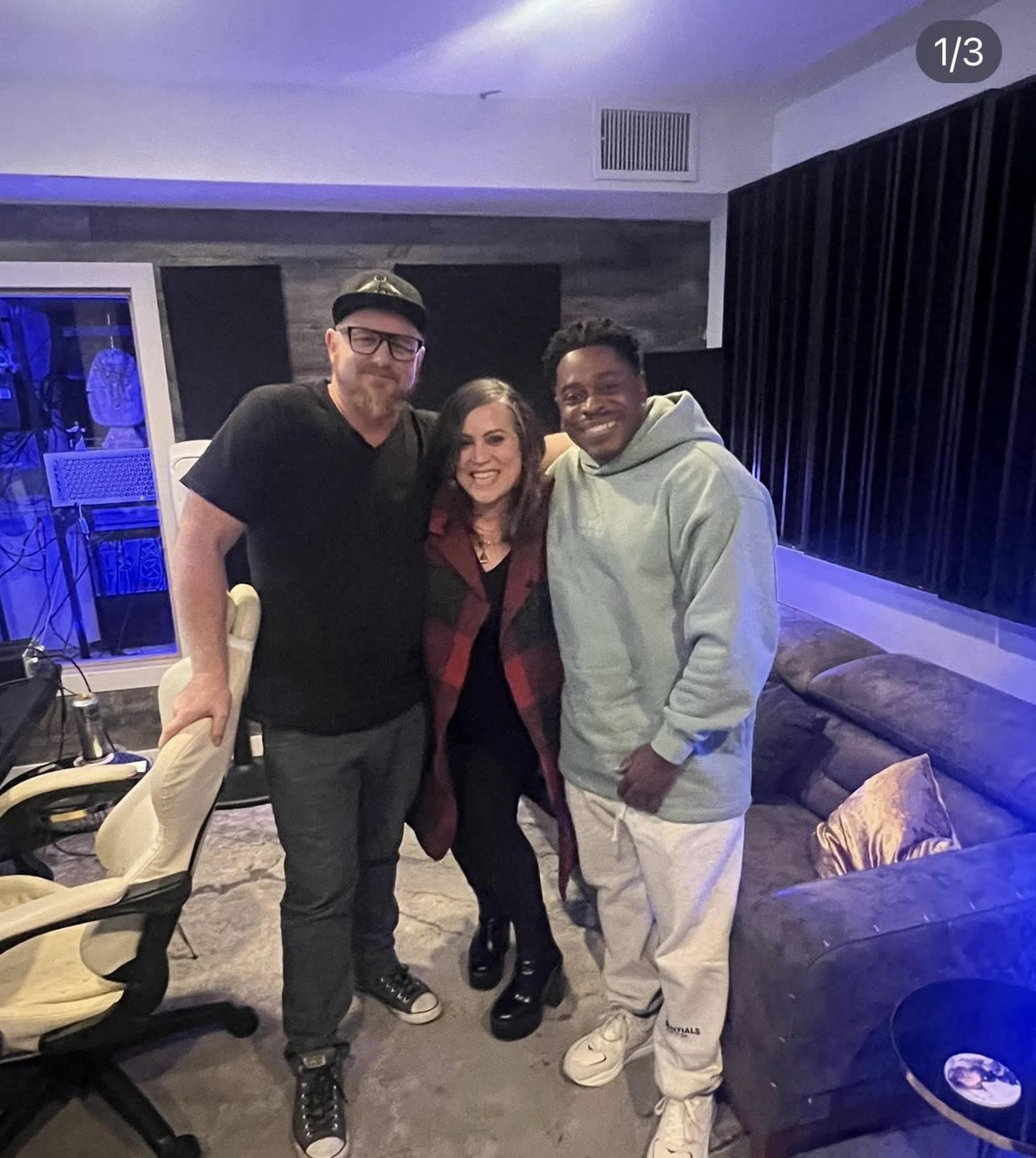 Three individuals pose together in a music studio with dark walls and a cozy seating area.