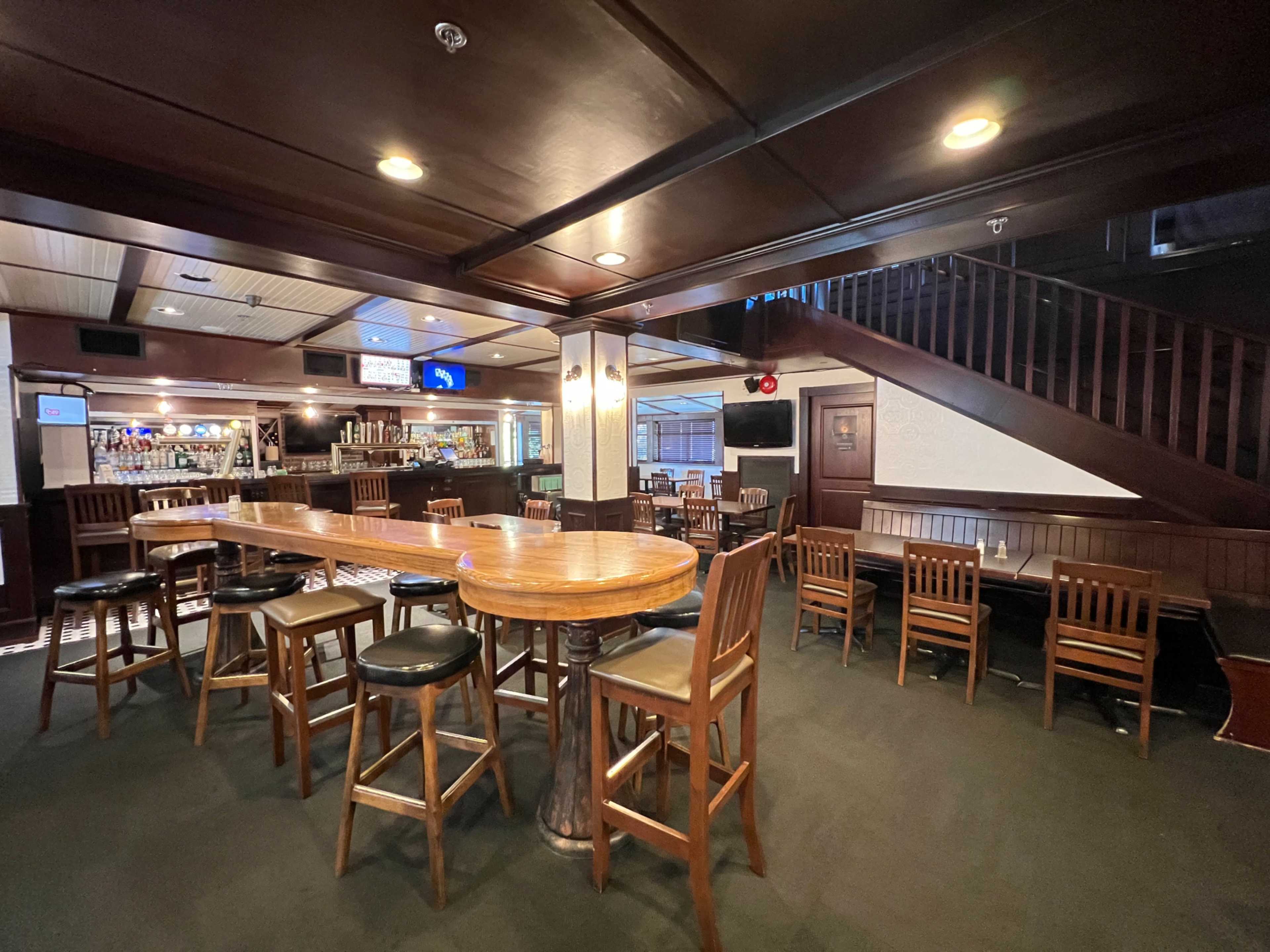 A spacious, dimly lit bar area with wooden furniture, a long table surrounded by high stools, and a staircase in the background.