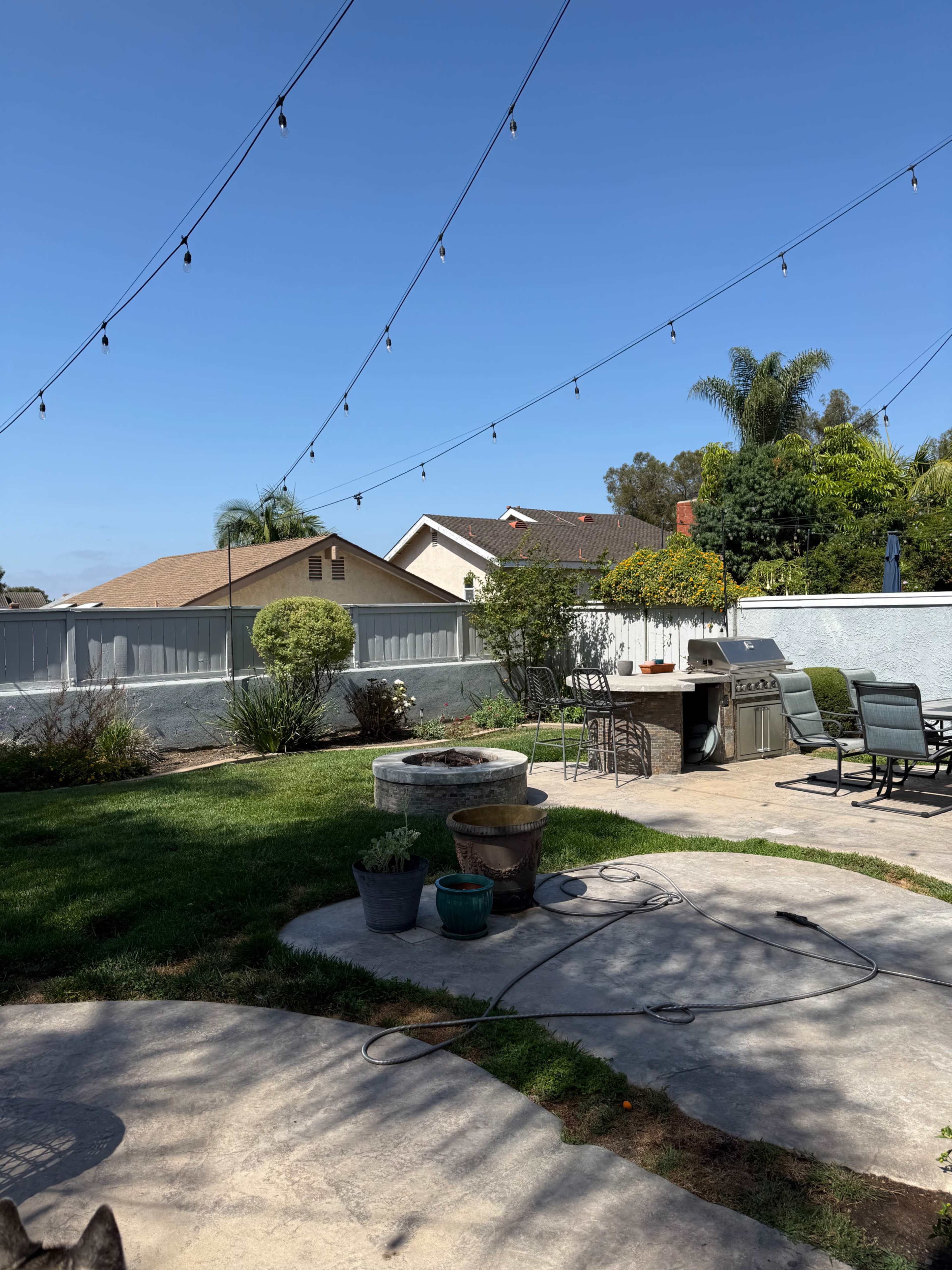 A backyard features a patio area with a fire pit, garden pots, and string lights overhead against a clear blue sky.