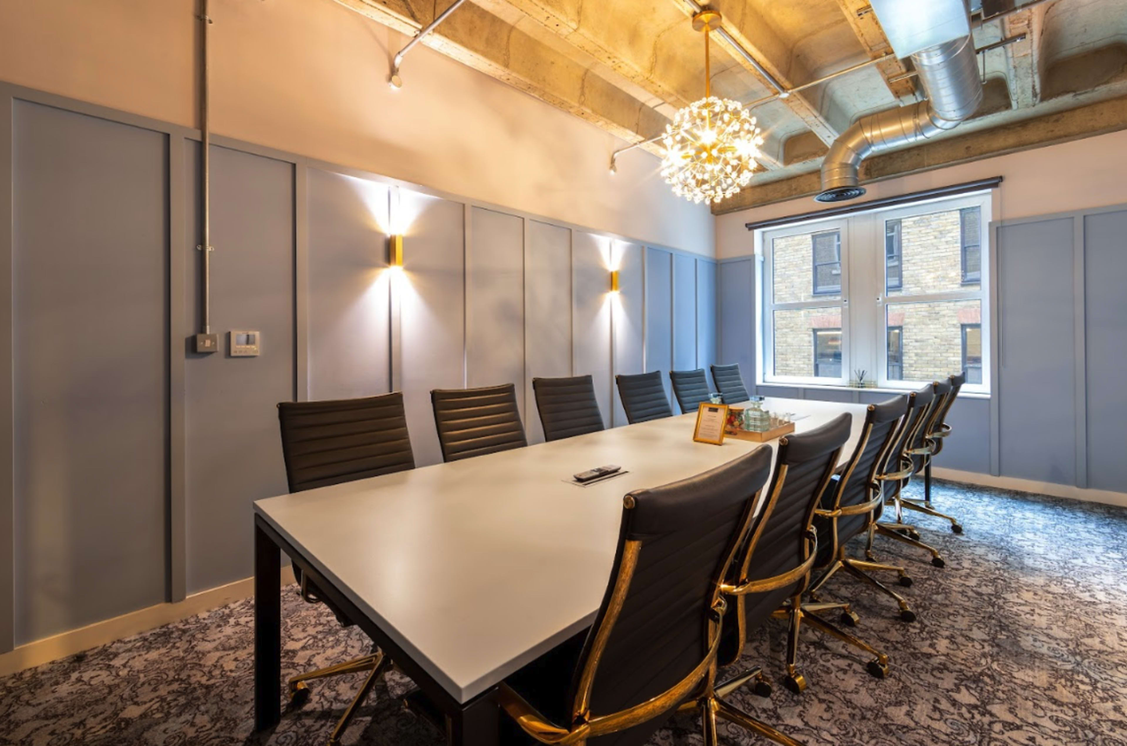 A modern conference room features a large table surrounded by black chairs, with decorative lighting and natural light streaming through a window.