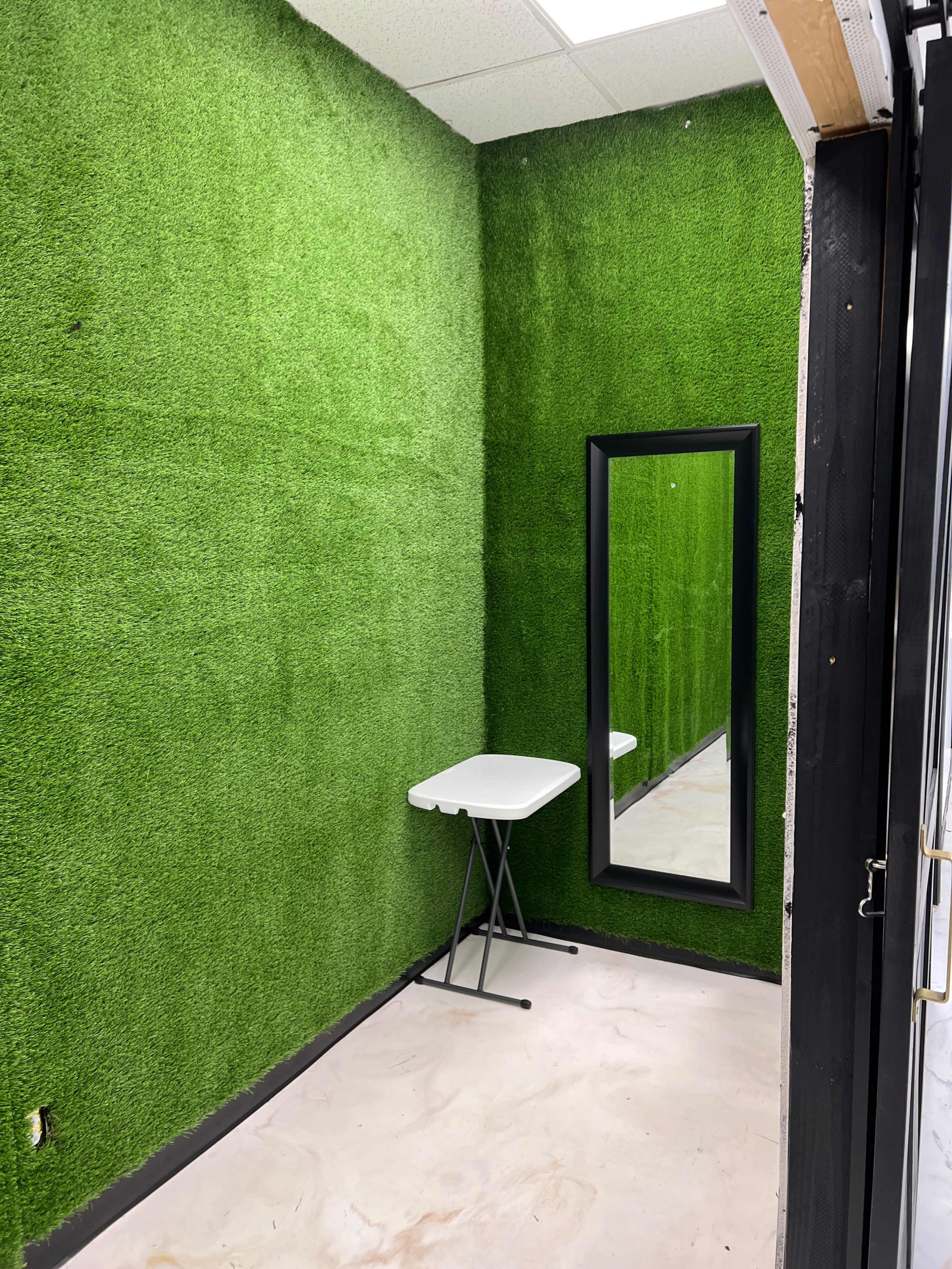 A small room with green artificial grass-covered walls, a folding table, and a tall mirror.