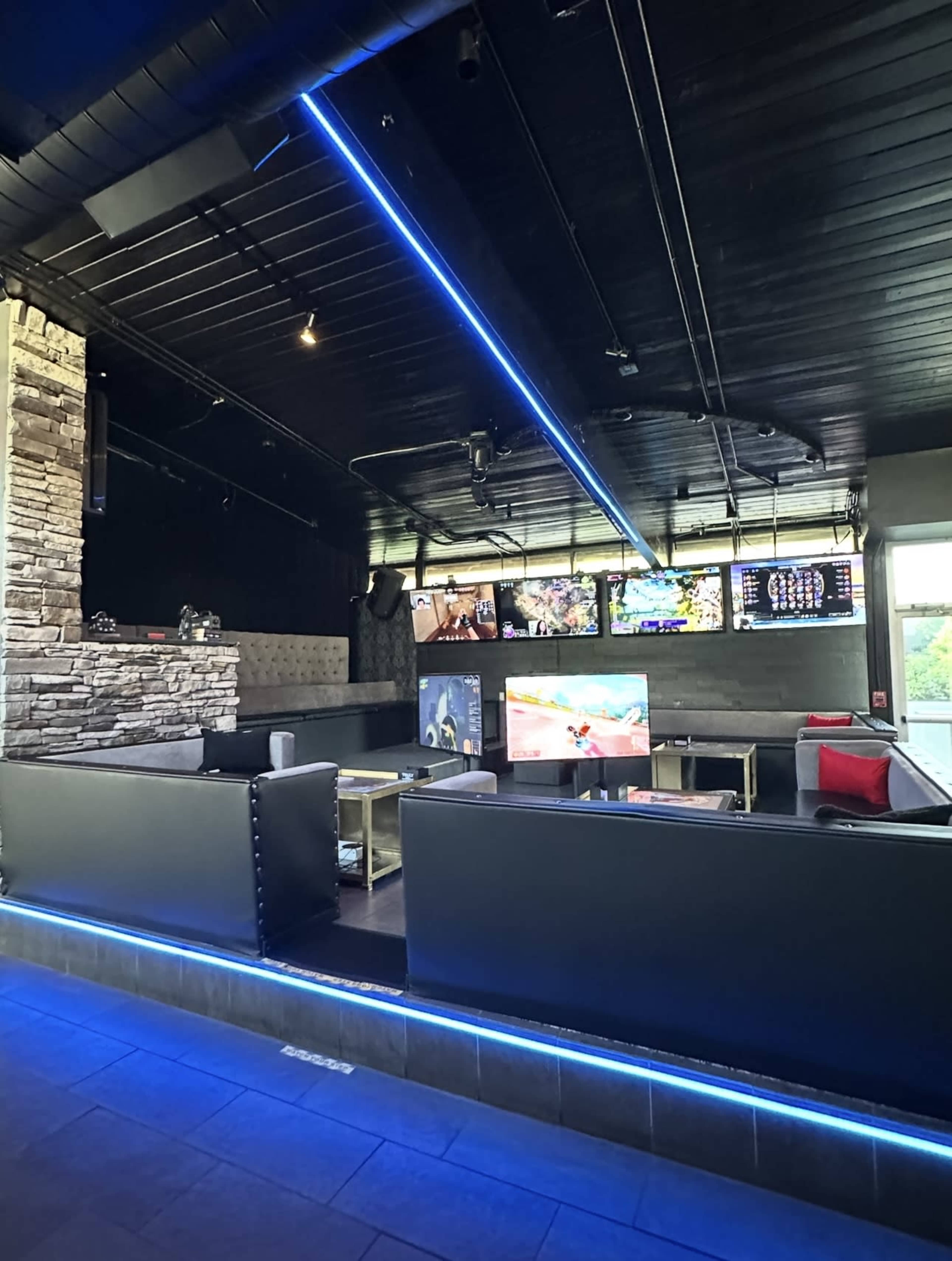 The image shows a modern lounge area featuring black furniture, multiple large screens displaying various content, and blue LED lighting along the walls.