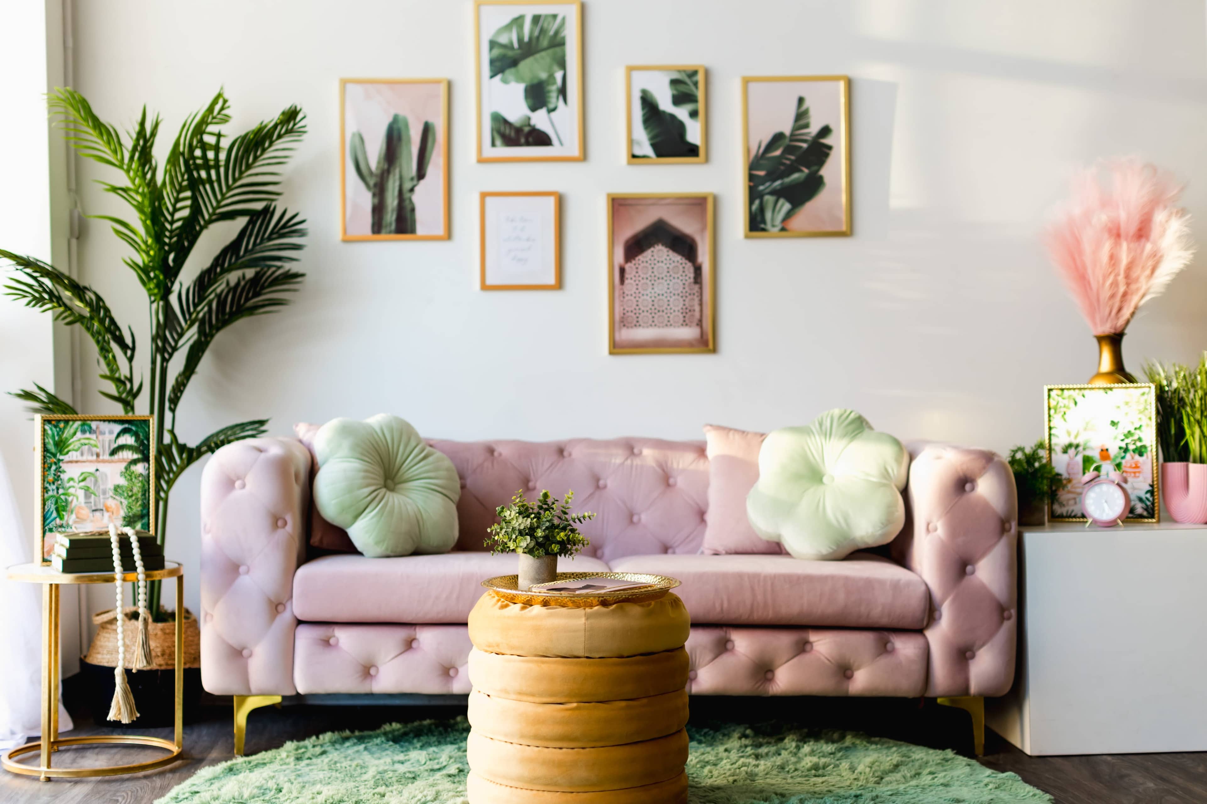 A pink tufted sofa with decorative pillows is positioned against a white wall adorned with framed botanical prints, accompanied by a round wooden coffee table and greenery.