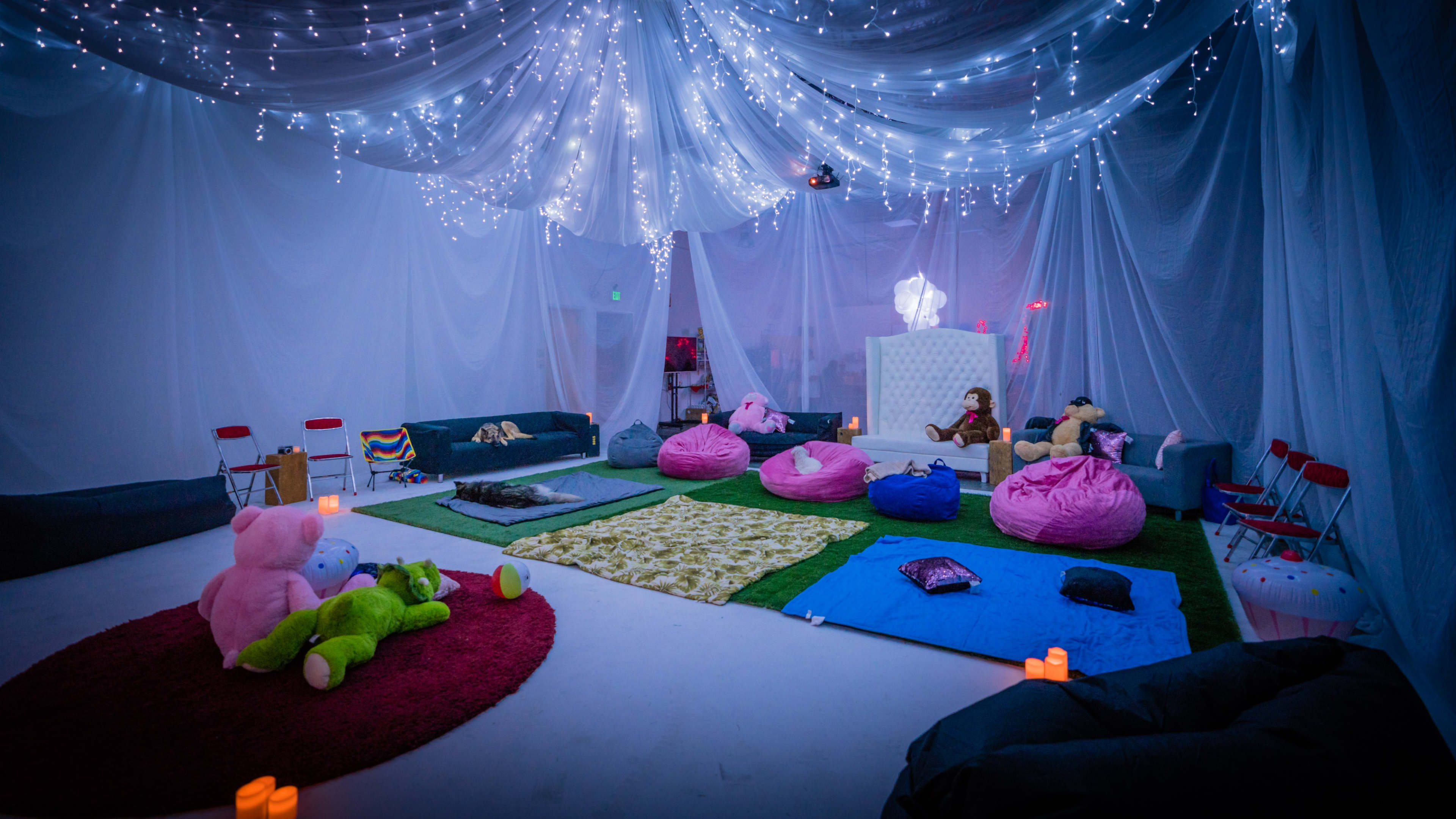 The scene shows a cozy indoor space decorated with draped fabric and fairy lights, featuring several colorful bean bags, rugs, and stuffed animals arranged on the floor.