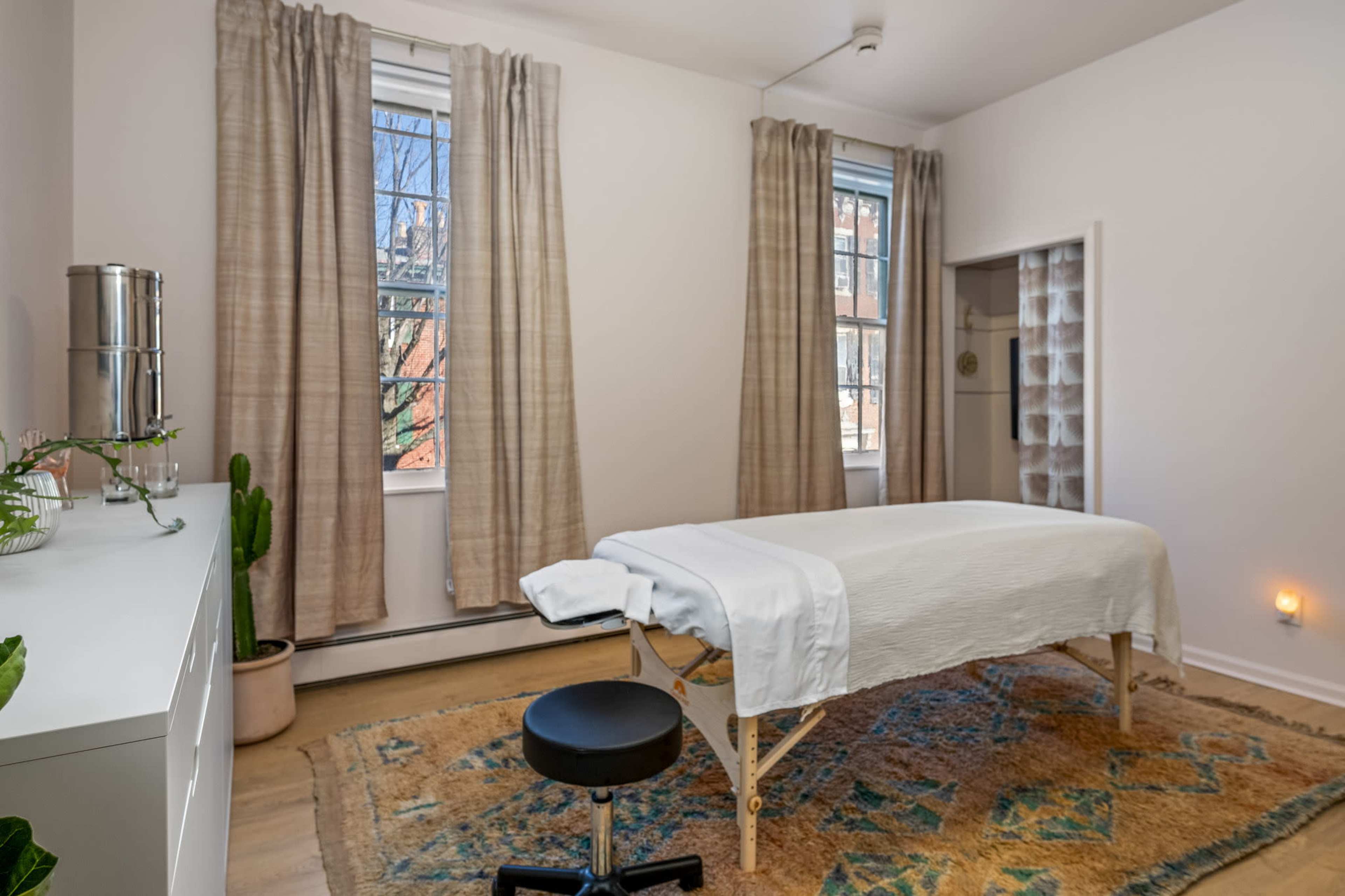 The image shows a massage therapy room with a treatment table, a black rolling stool, and large windows with sheer curtains.