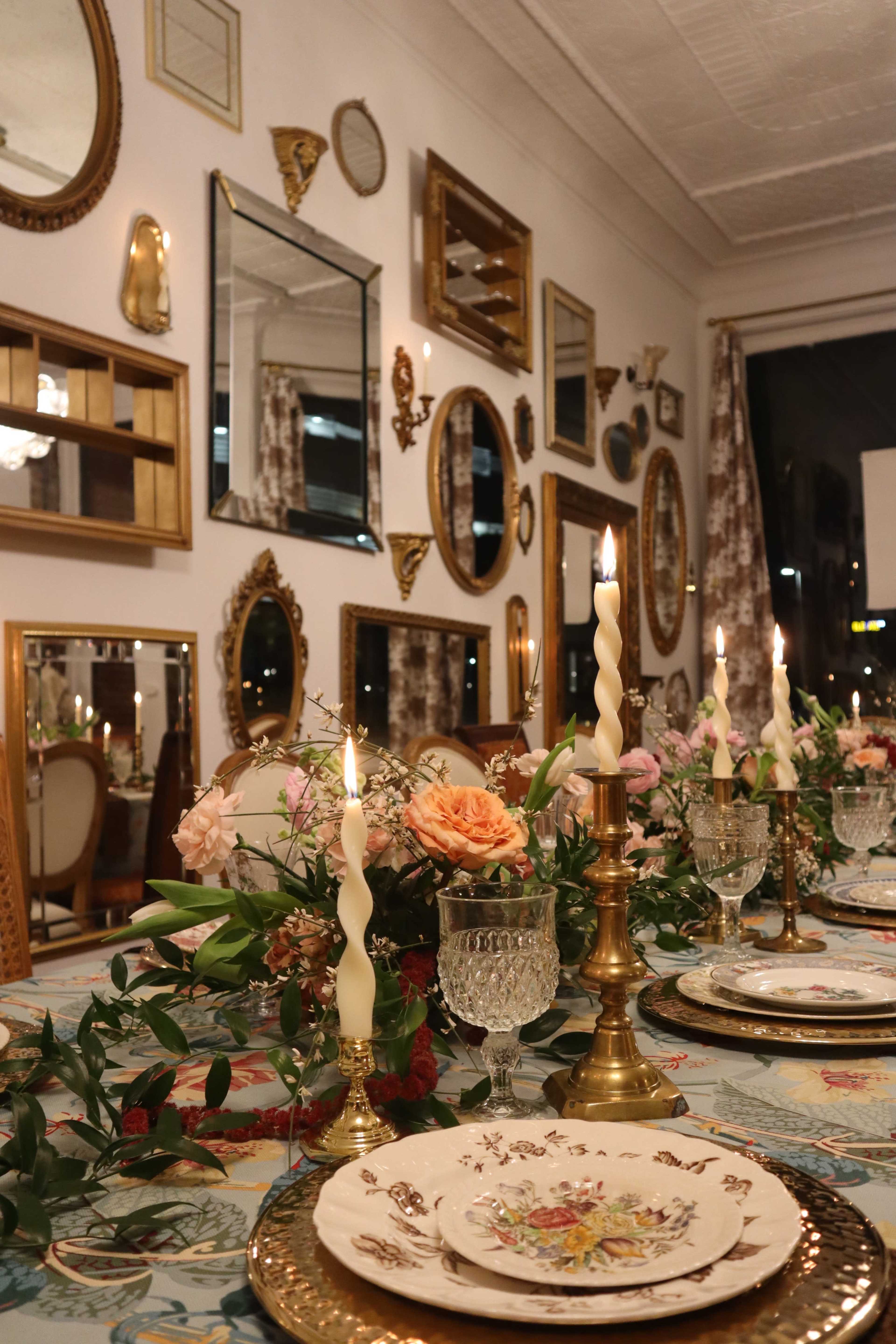 A beautifully set dining table with floral arrangements, vintage tableware, and lit candles is surrounded by an array of decorative mirrors on the walls.