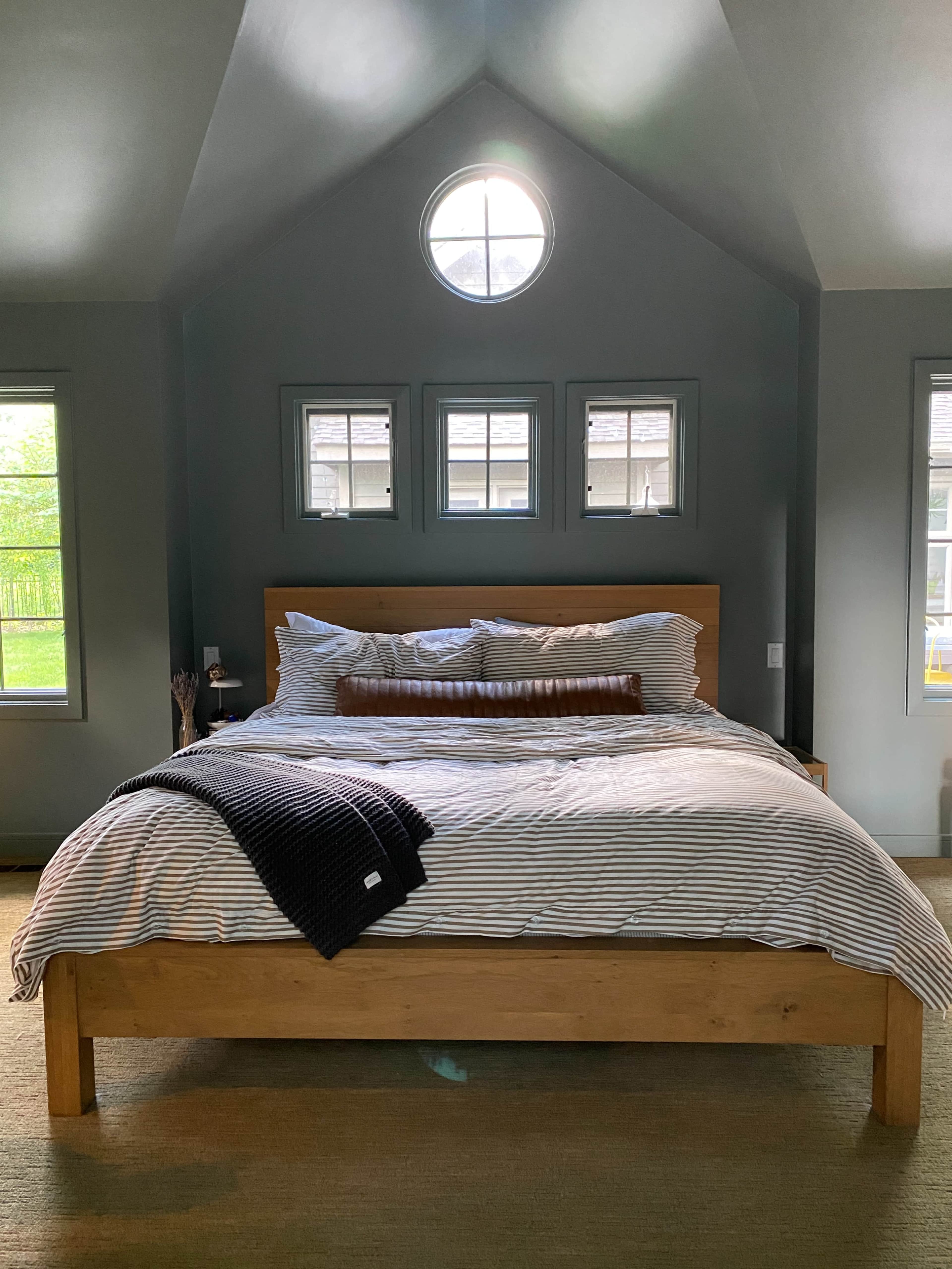 A wooden bed with striped bedding is centered in a room featuring a circular window and several smaller windows on the walls.