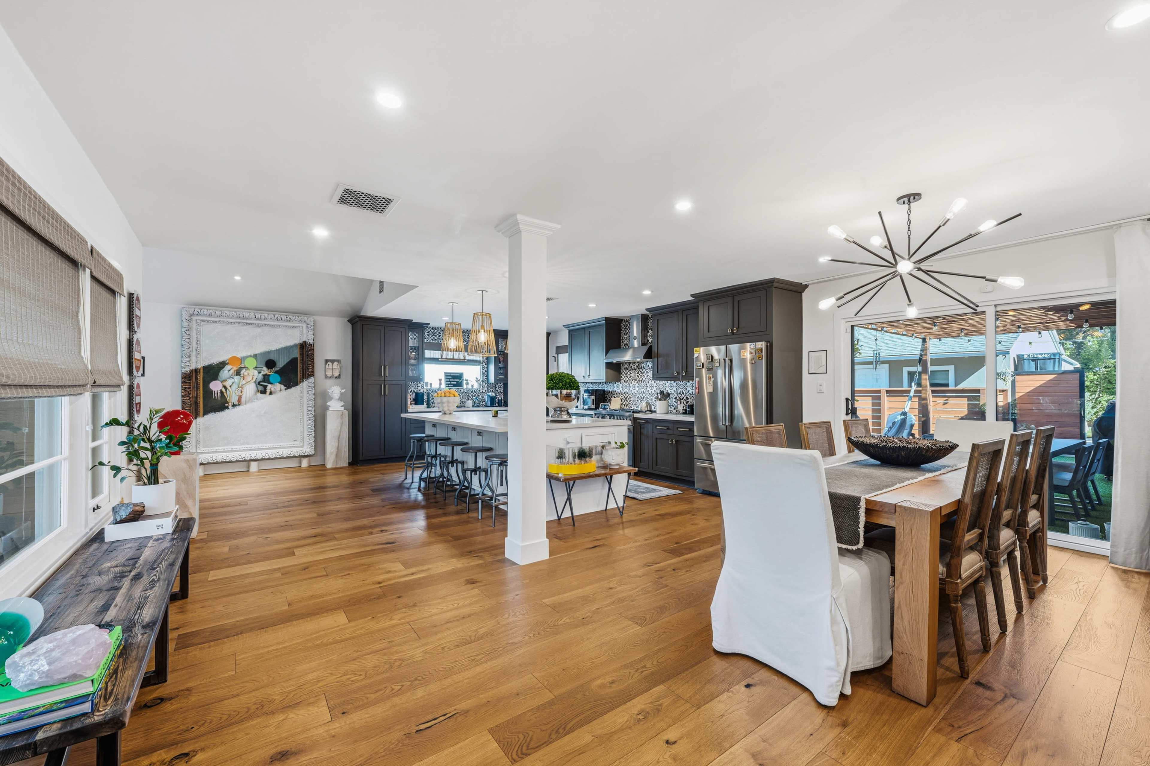 The image shows a modern open-concept kitchen and dining area with hardwood floors, featuring dark cabinetry, a large dining table, and an assortment of stylish lighting fixtures.