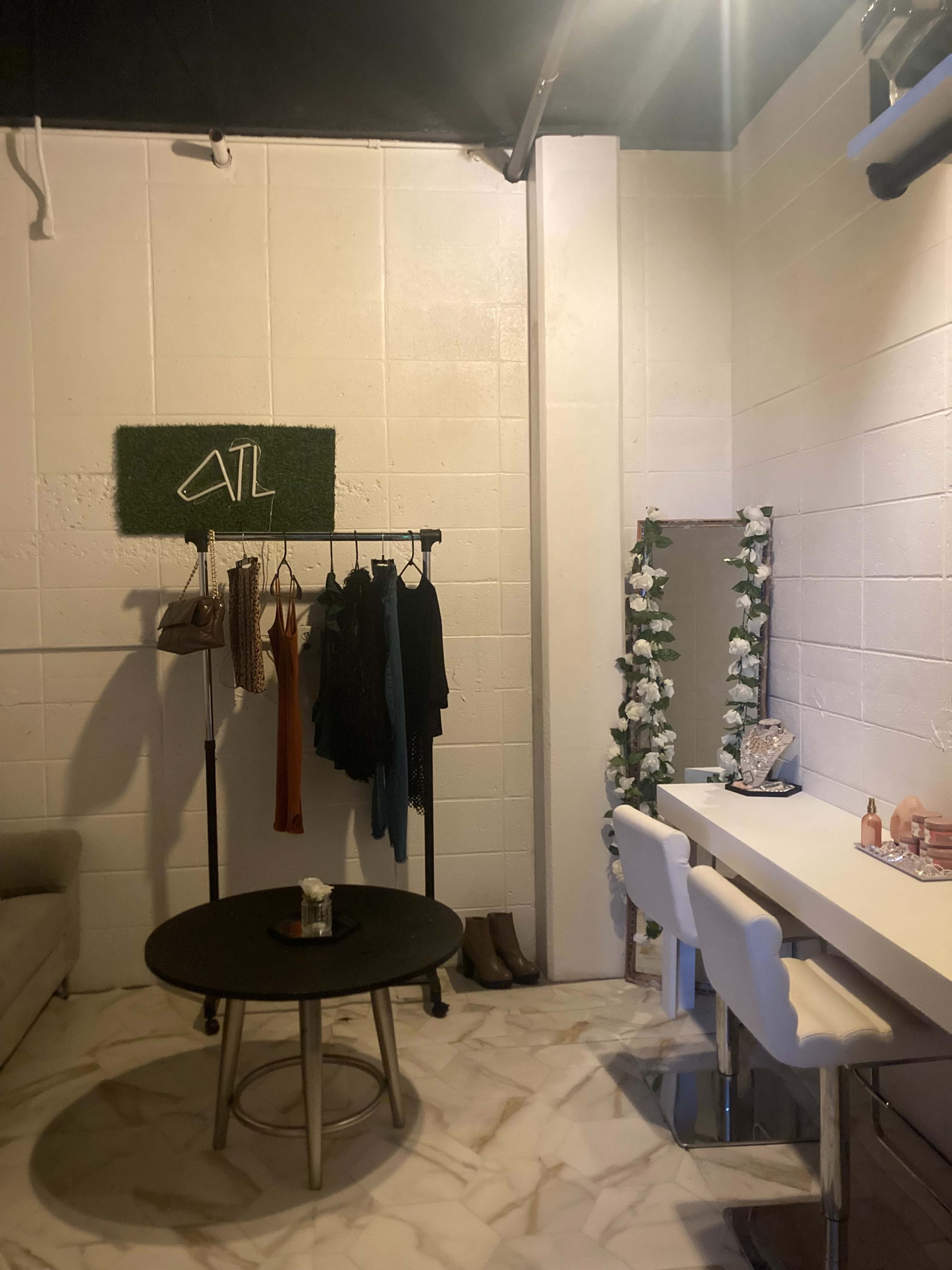 A small room with a clothing rack displaying several garments, a round table in the center, and a vanity area with white stools and a mirror surrounded by flowers.