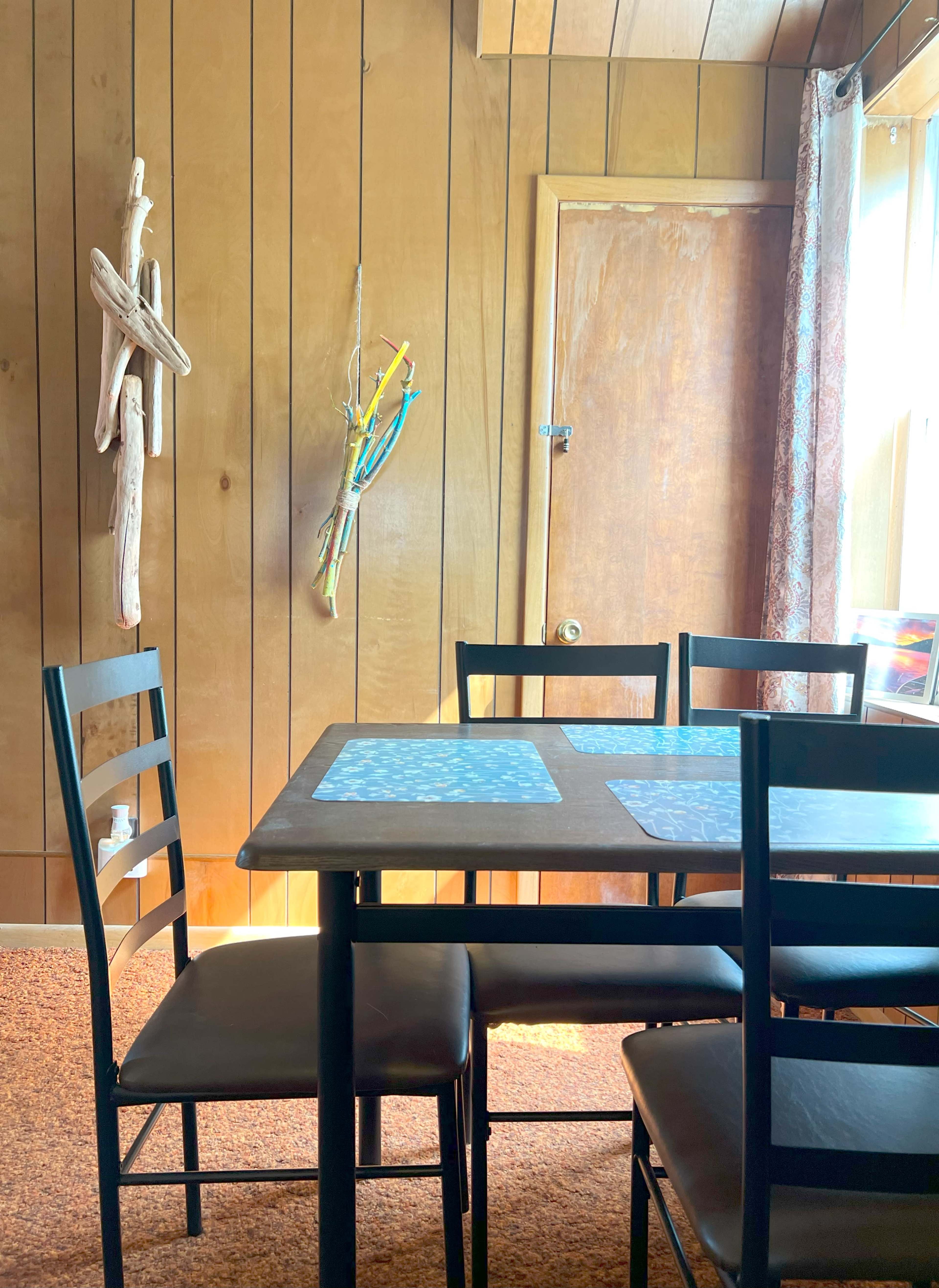 A simple dining area features a table with blue placemats and four black chairs, set against a wooden-paneled wall.