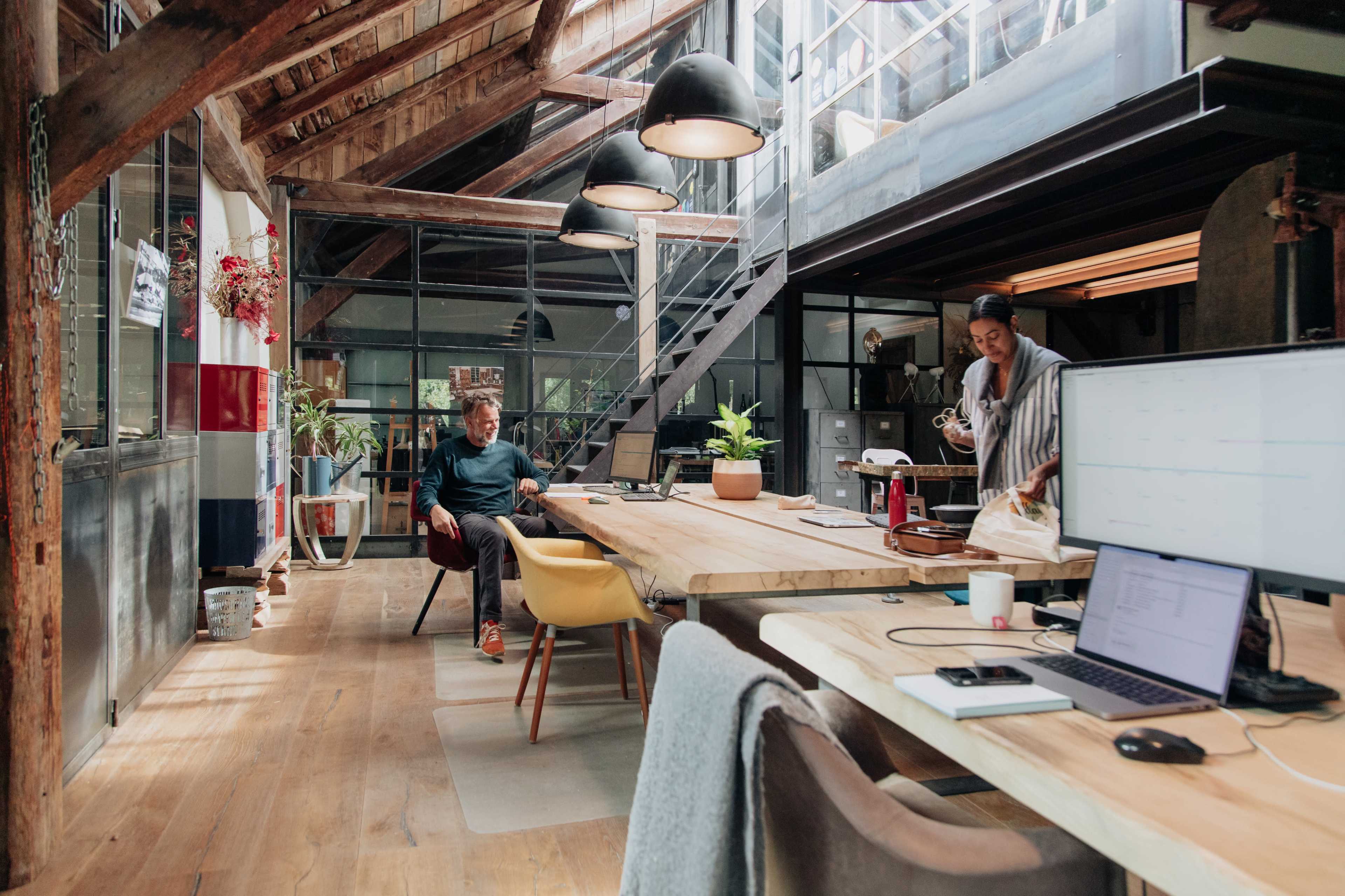 A bright, industrial-style office features wooden beams, large windows, and a long table with a person working at a computer while another prepares coffee.