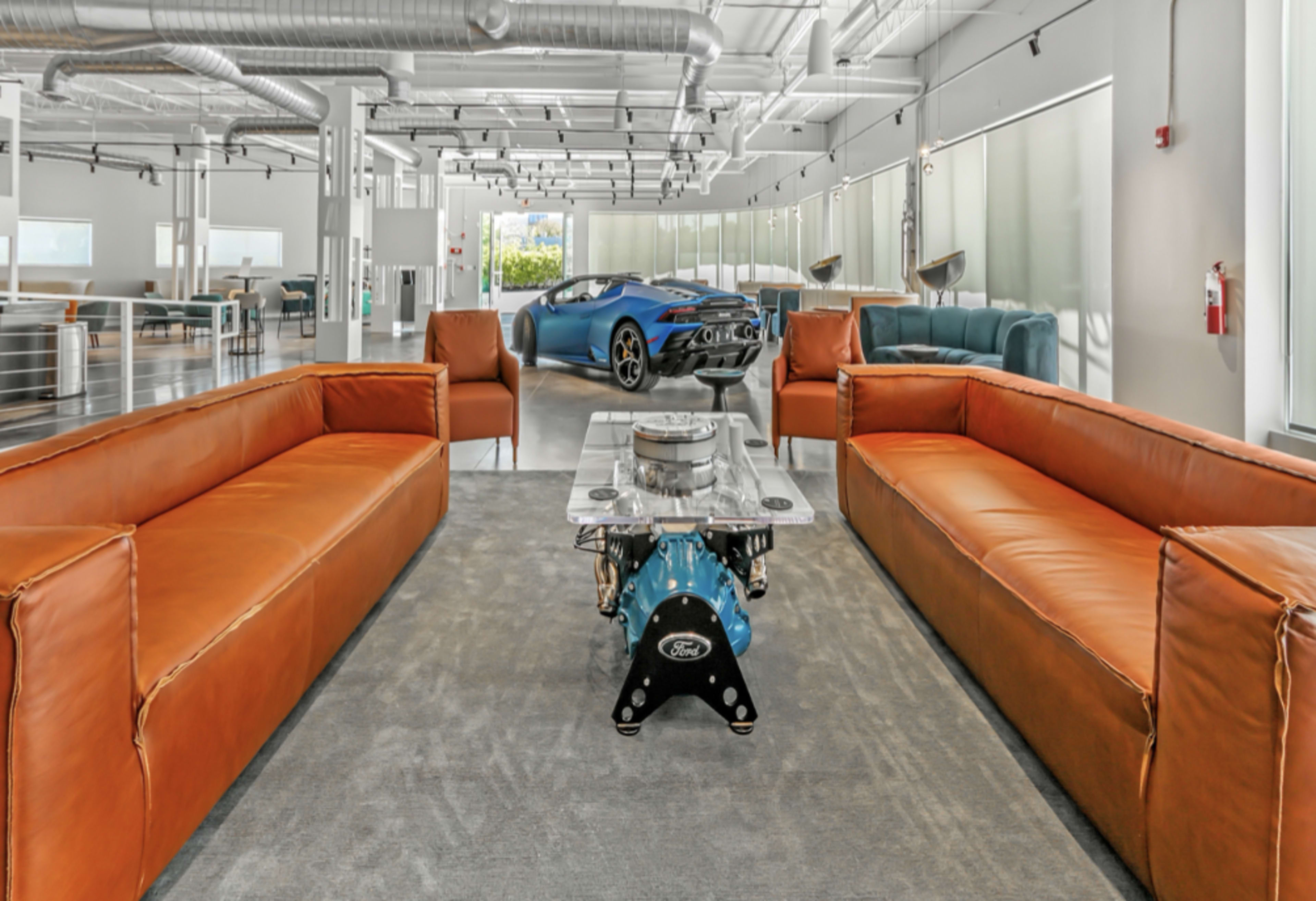 A modern lounge area with two leather sofas and a glass coffee table, featuring a blue sports car visible in the background.