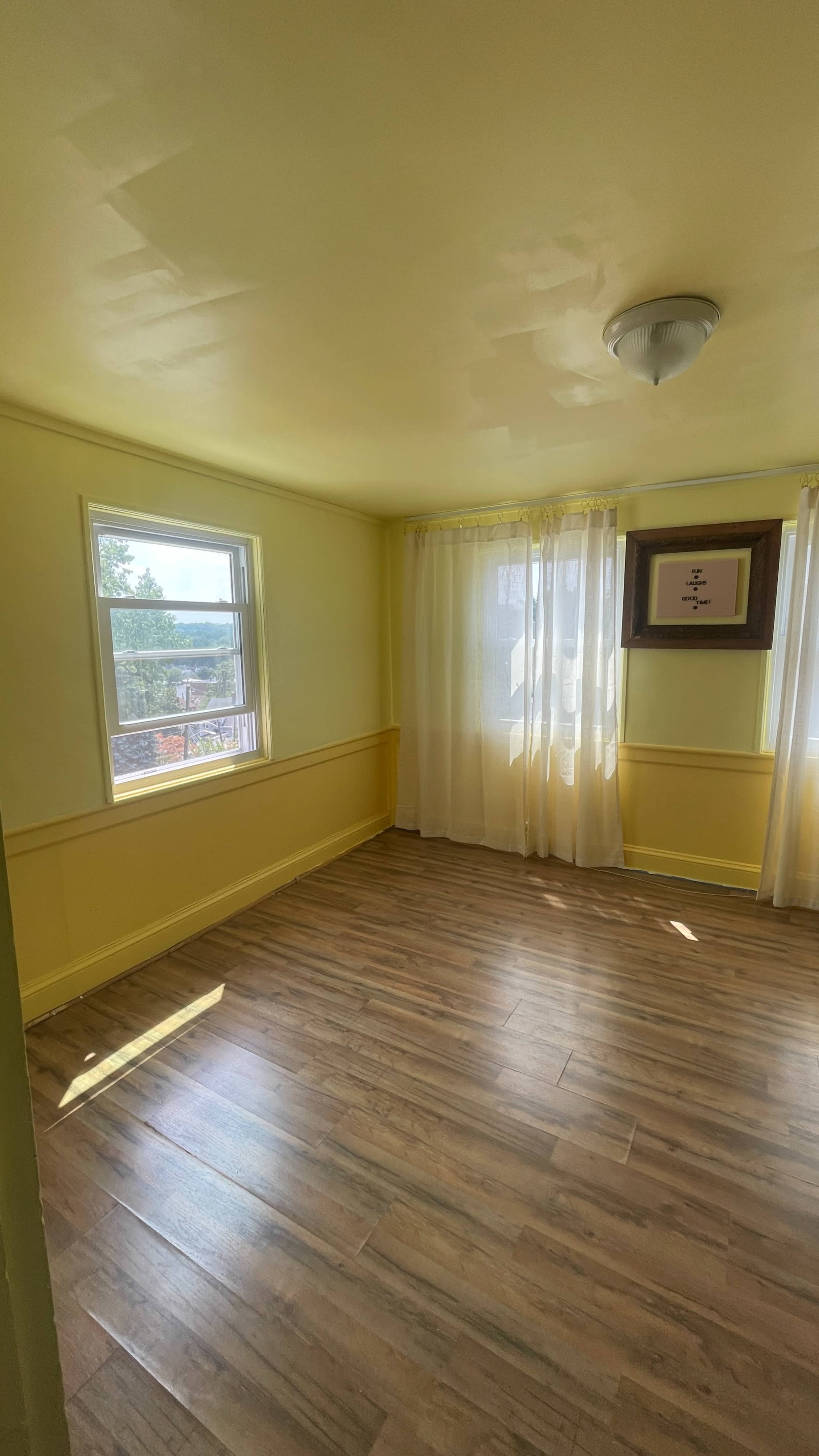 The room features yellow walls, two windows adorned with sheer curtains, and a wooden floor.