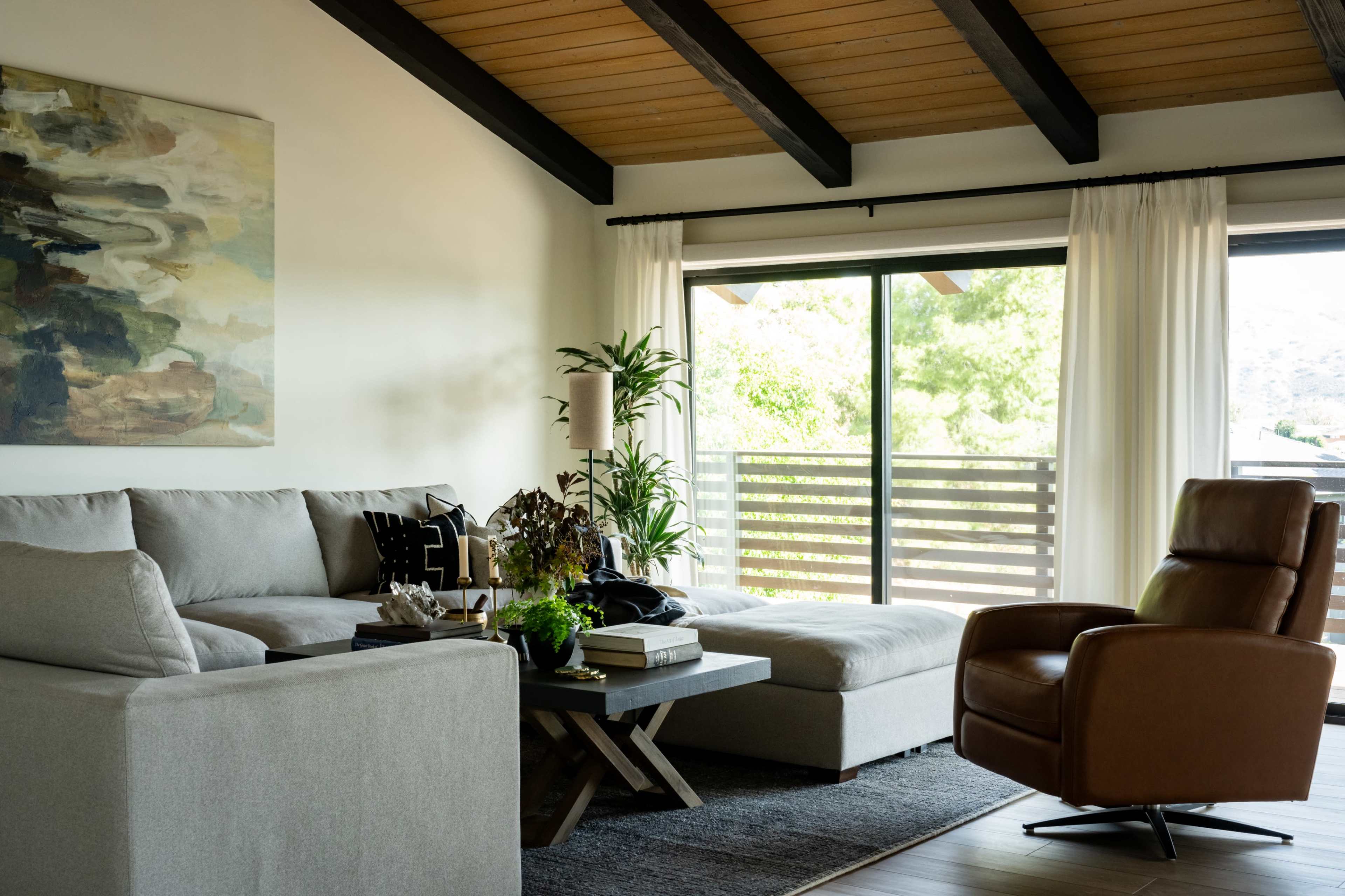 A spacious living room features a sectional gray sofa, a brown recliner, a coffee table with decor items, and large windows framed by white curtains.