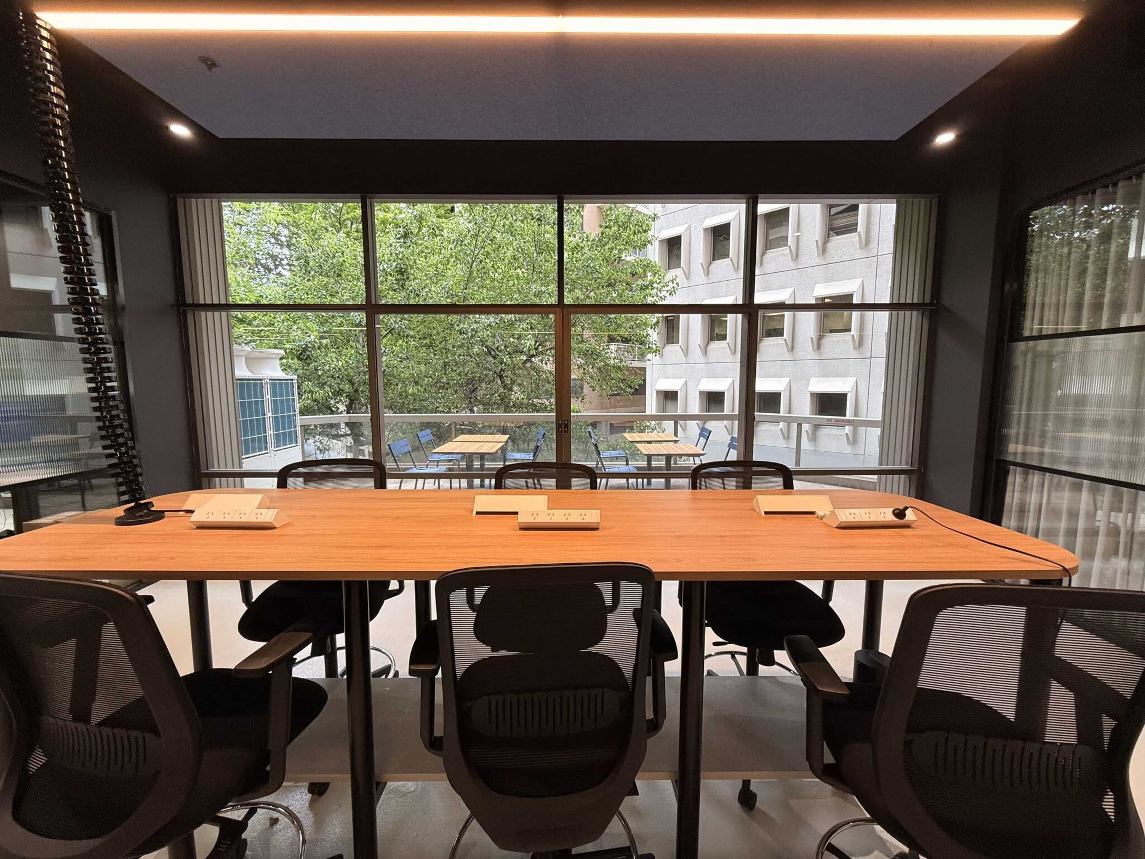 A modern conference room features a long wooden table surrounded by ergonomic chairs, with large windows showcasing an outdoor seating area and greenery.