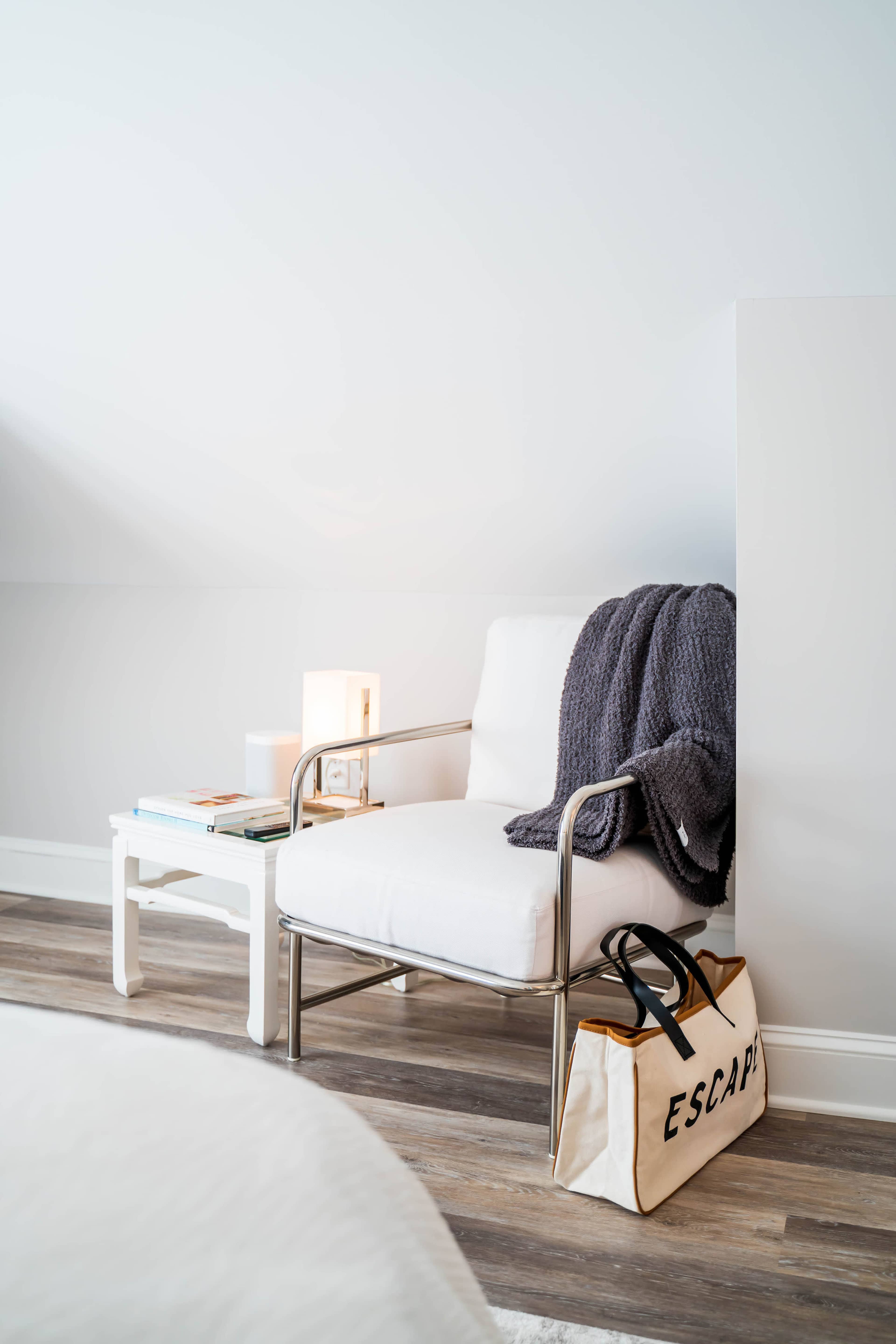 A modern white chair with a gray blanket draped over it sits next to a small table and a bag labeled "ESCAPE" on the floor.