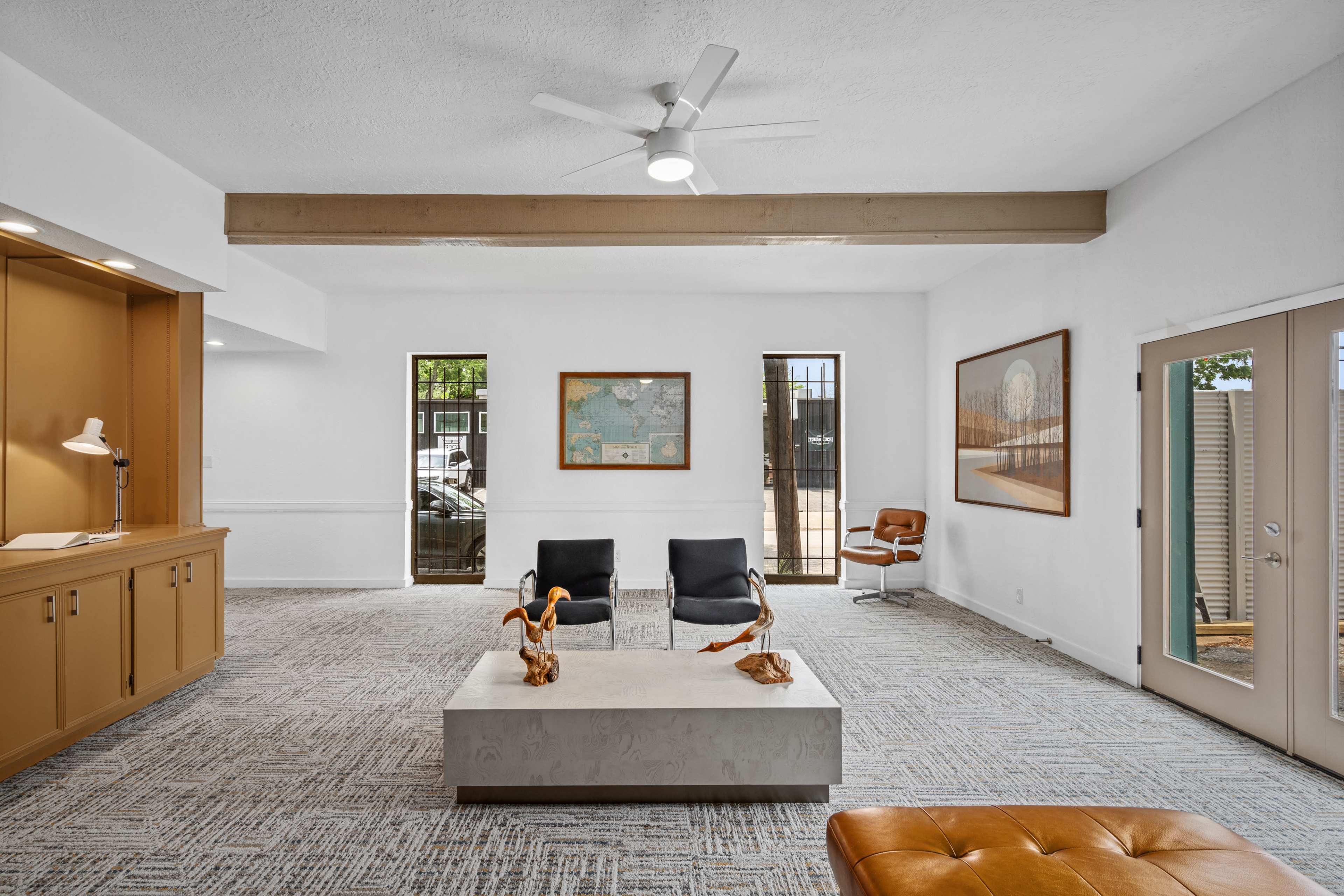 The image shows a bright, modern interior space featuring two black chairs, a light-colored coffee table, and a brown chair near large windows.