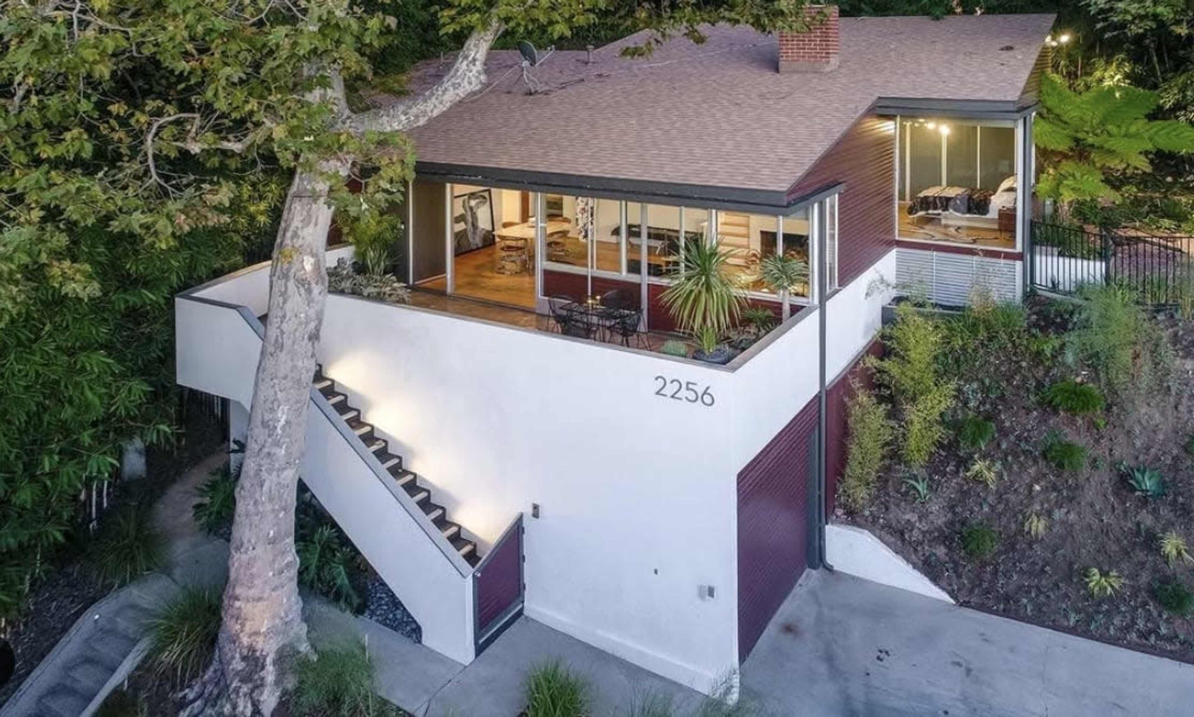 The image shows a modern two-story house with large windows and a prominent staircase leading to the entrance, surrounded by greenery.