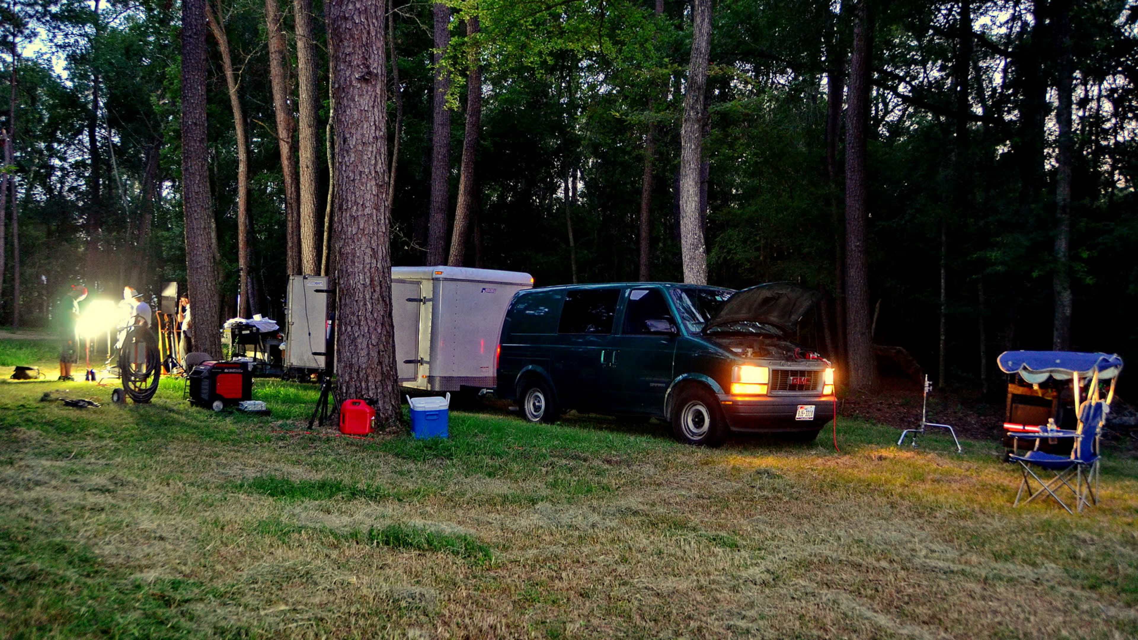 A green van with its hood open is parked next to a trailer in a wooded area, illuminated by bright lights along the side.
