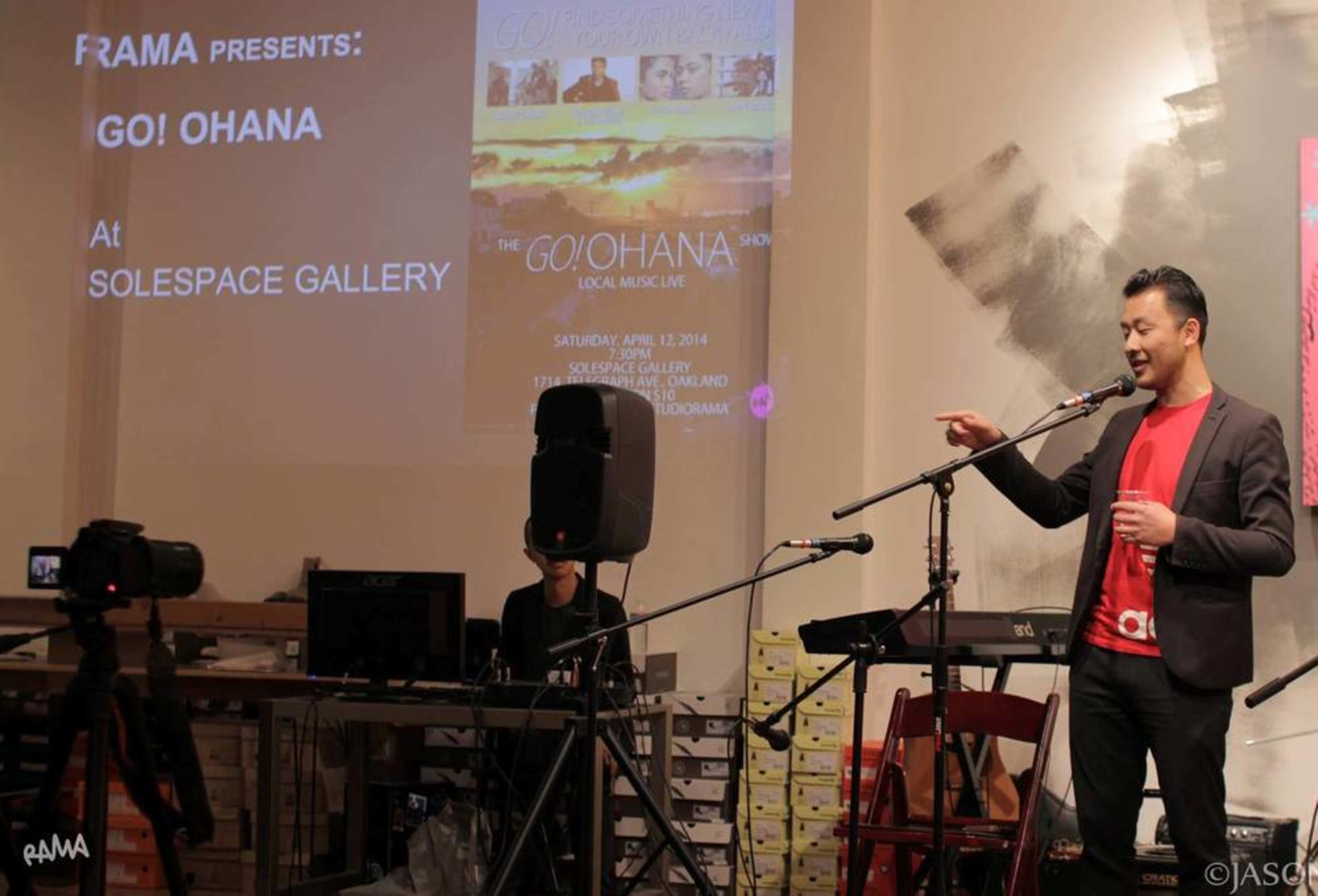 A man stands on stage speaking into a microphone, while a promotional poster for the Go! Ohana Show is displayed in the background.