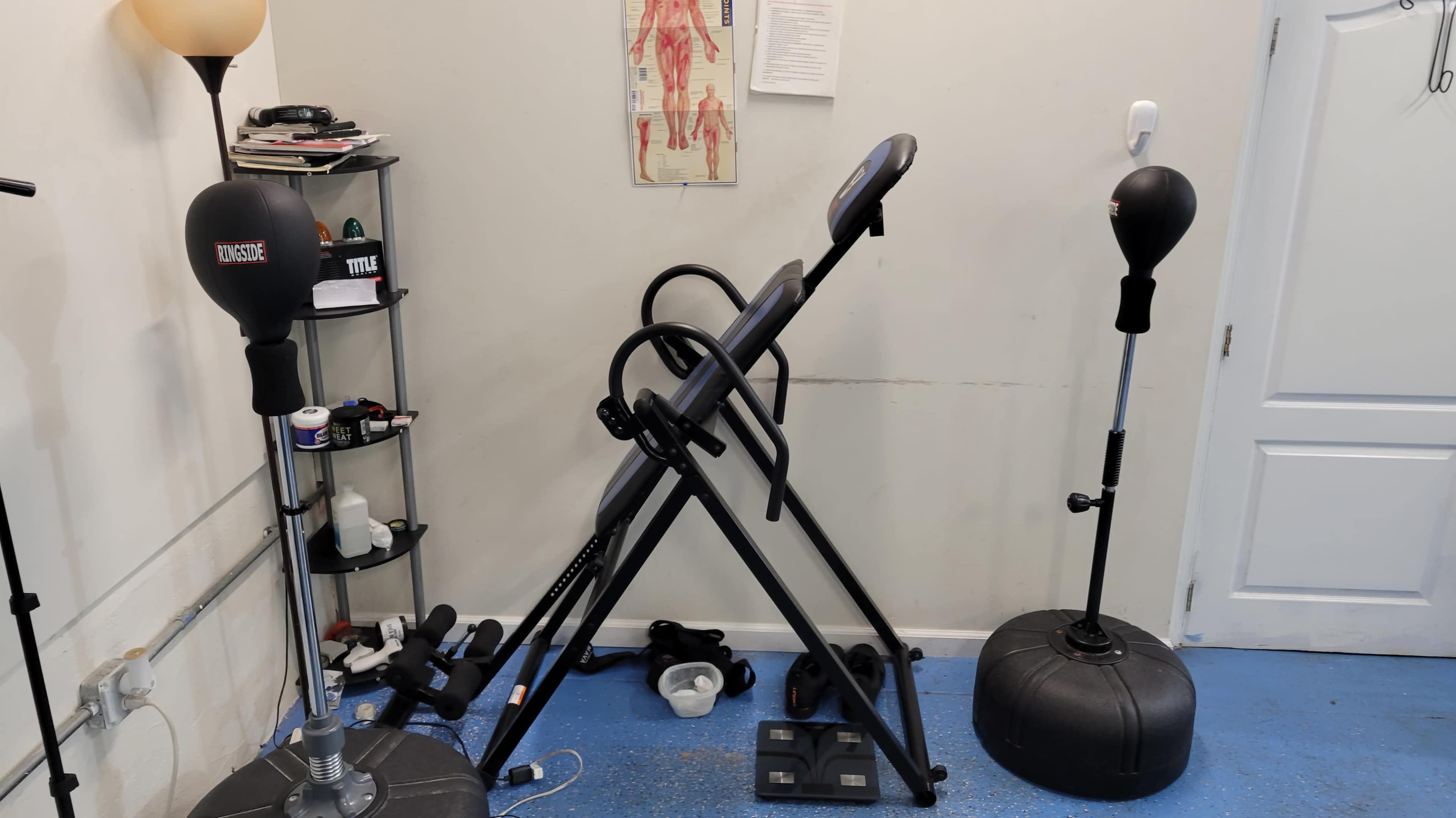 The image shows a small workout area featuring an inversion table, a punching bag on a stand, and various fitness accessories on a shelving unit.
