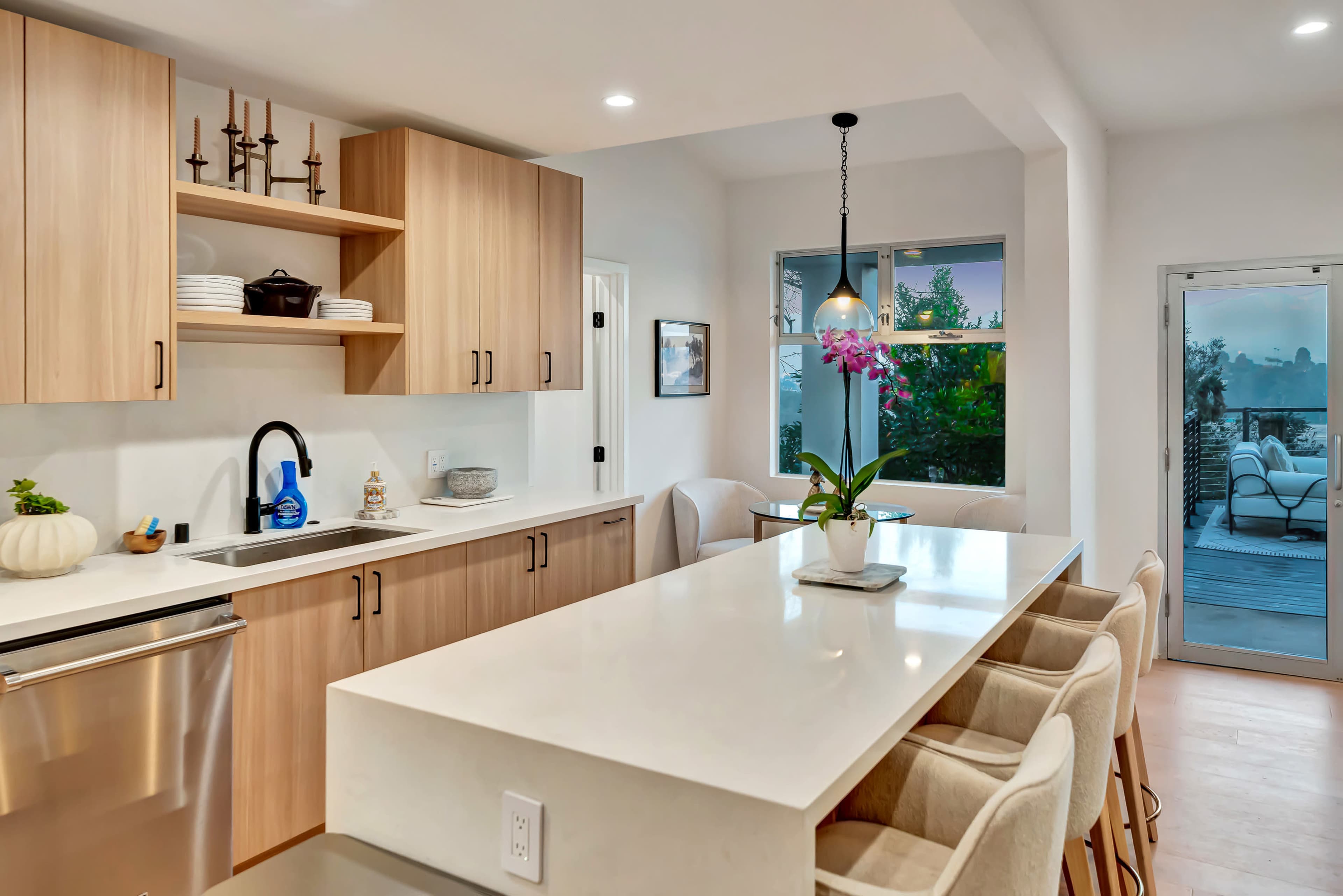 The modern kitchen features light wood cabinetry, a large island with seating, and a view of an outdoor area through a sliding glass door.