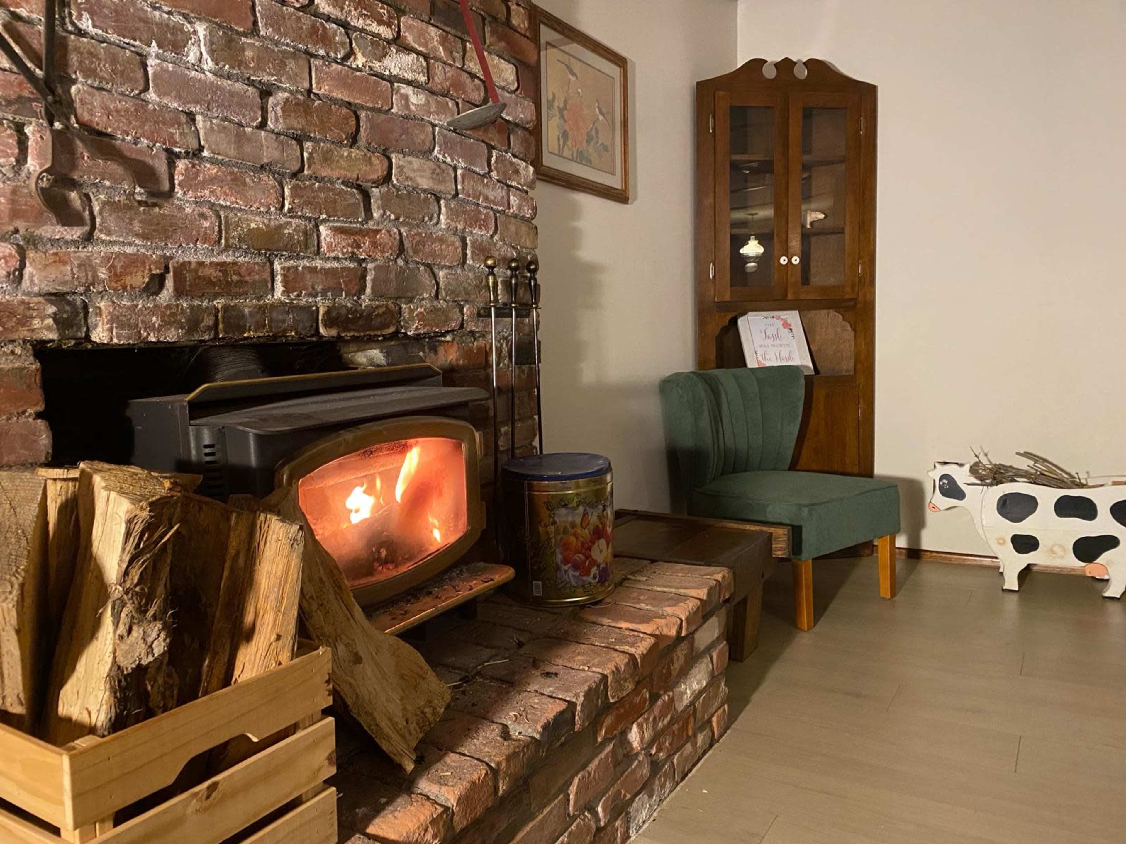 A brick fireplace with a wood stove is flanked by a green chair and a wooden cabinet in a cozy room.