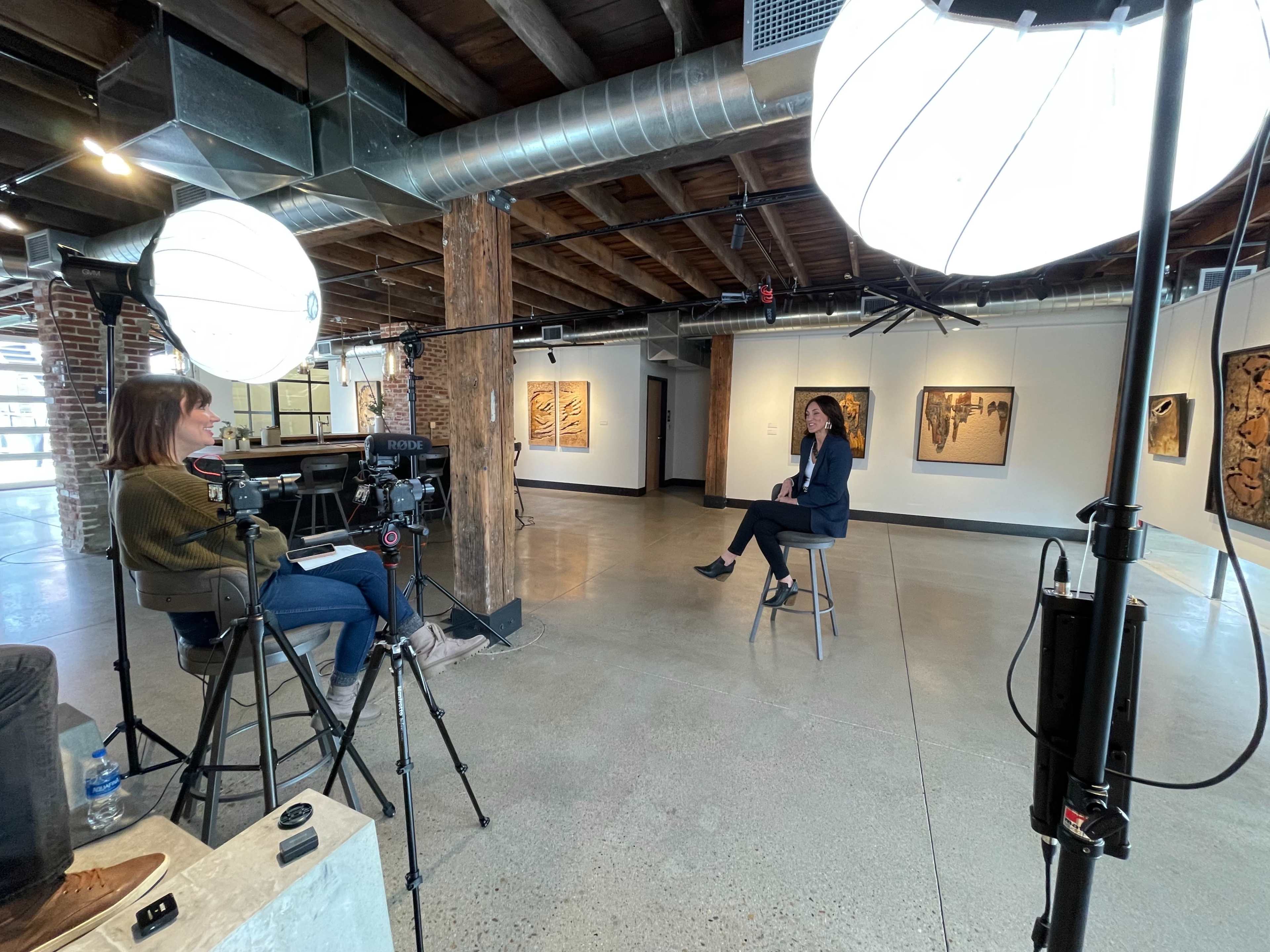 A woman is being interviewed on a stool in a gallery, with two large lights and cameras set up in a well-lit space featuring artwork on the walls.