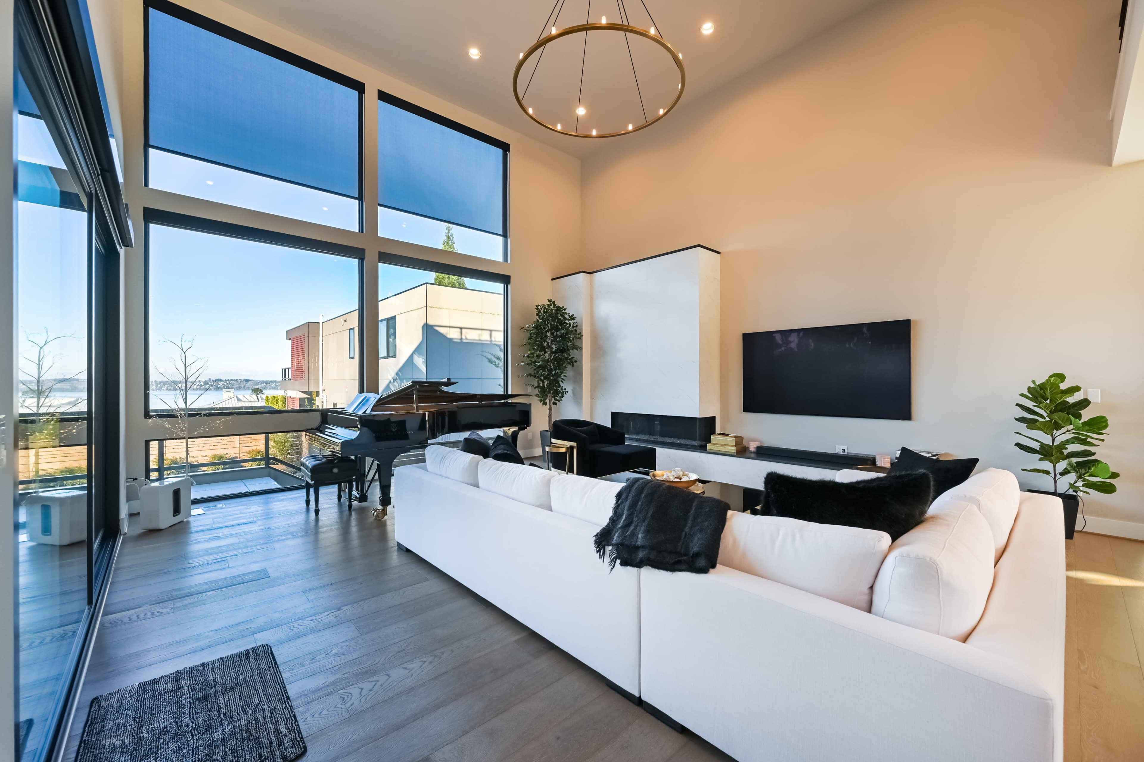A modern living room features a white sofa, a grand piano, and large windows that provide a view of the outdoor landscape.