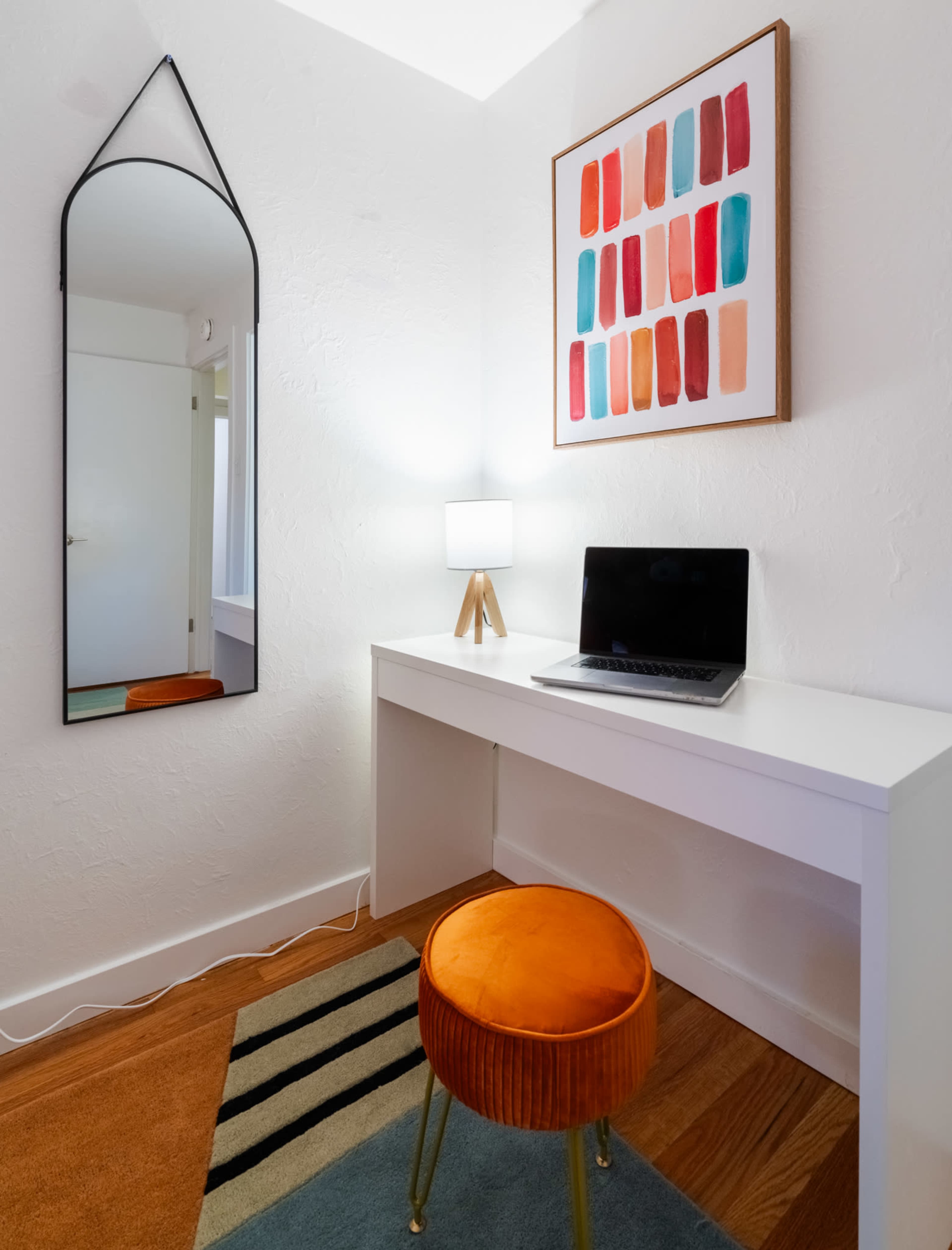 A small workspace features a white desk with a laptop, an orange stool, a lamp, a large mirror, and a colorful art piece on the wall.