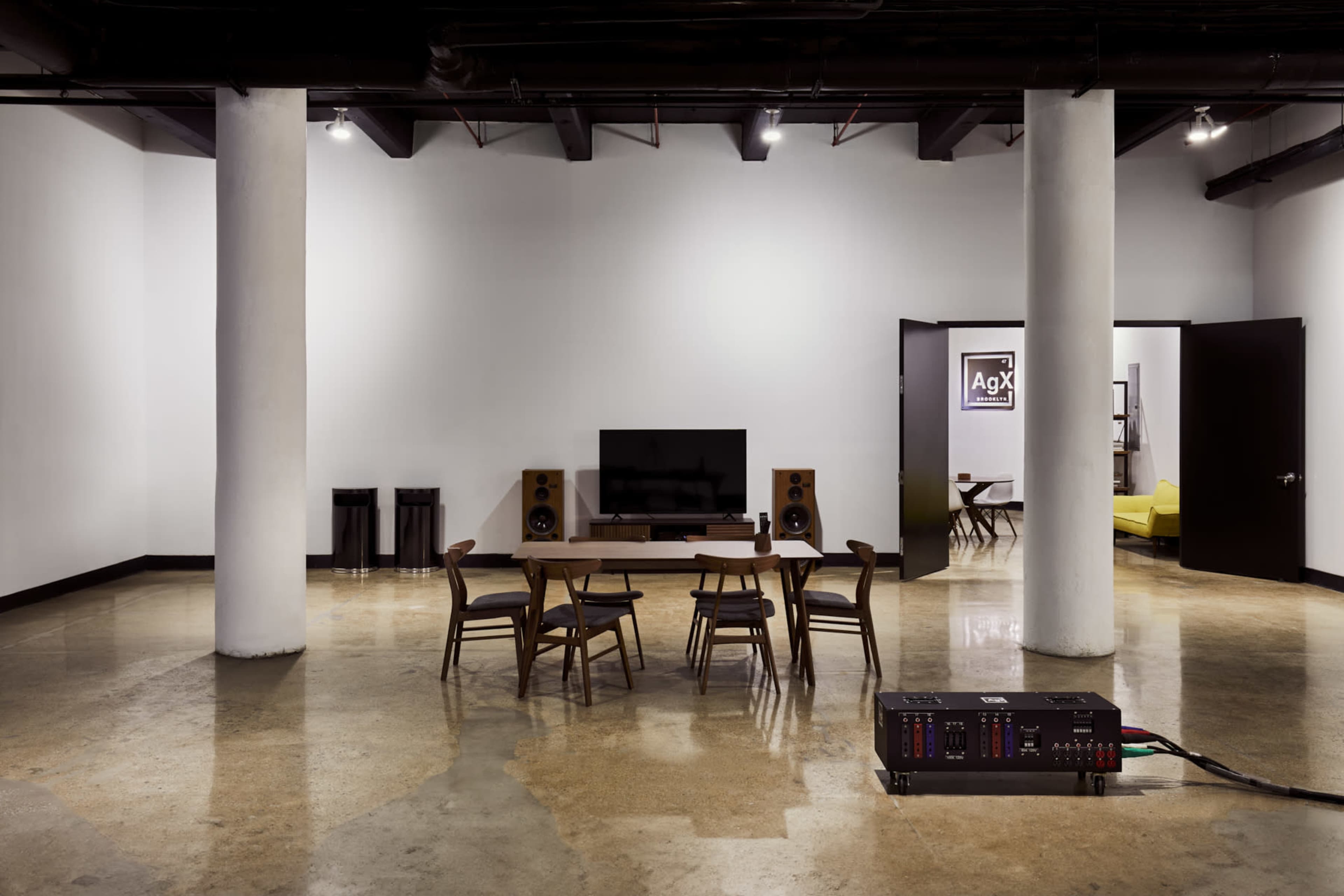 The image depicts a spacious, modern room with polished concrete flooring, featuring a wooden dining table surrounded by four chairs, a flat-screen TV mounted on the wall, and audio equipment on either side.