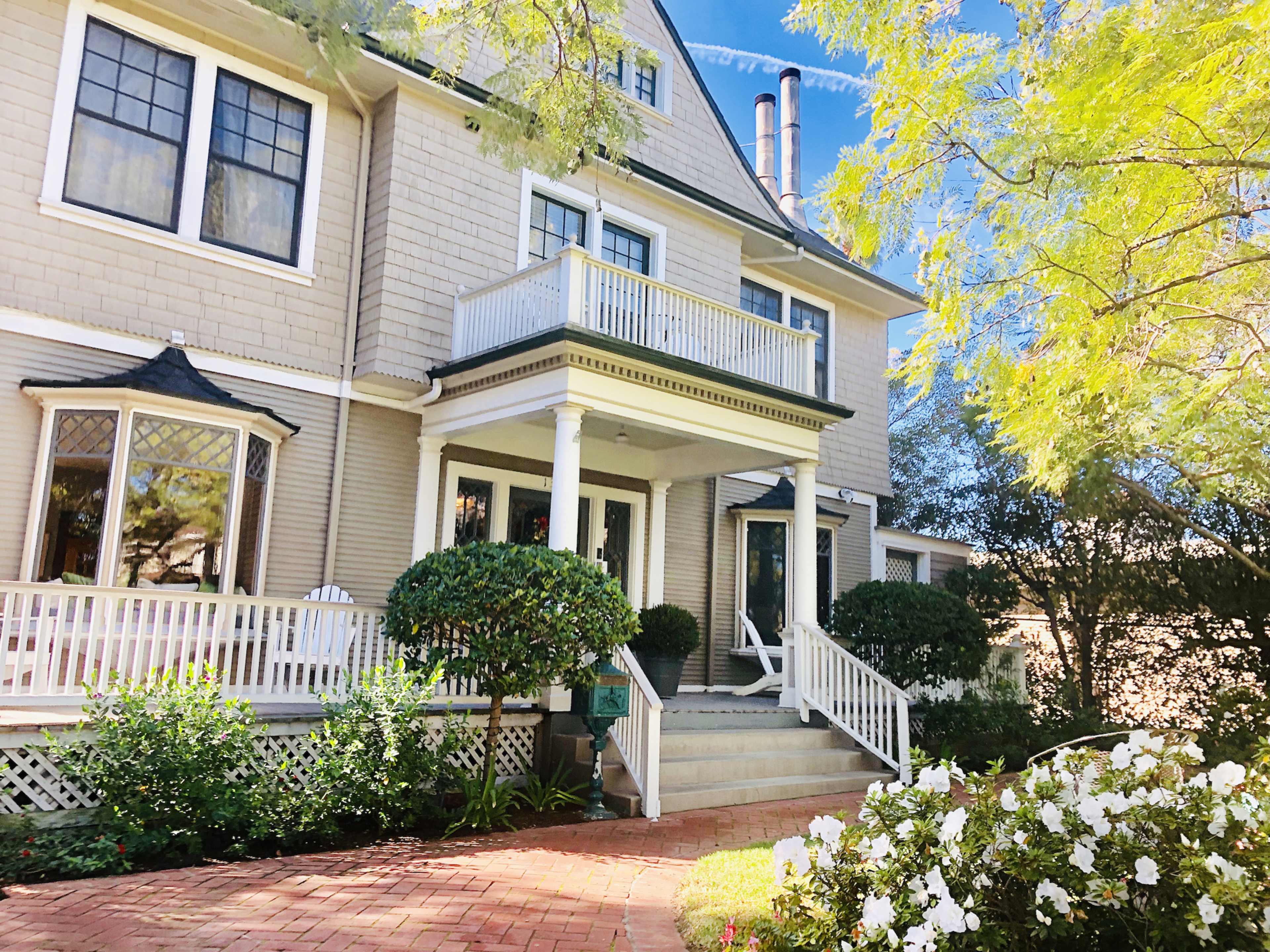 A two-story house with a wraparound porch, surrounded by well-maintained gardens featuring white flowers and shrubs.