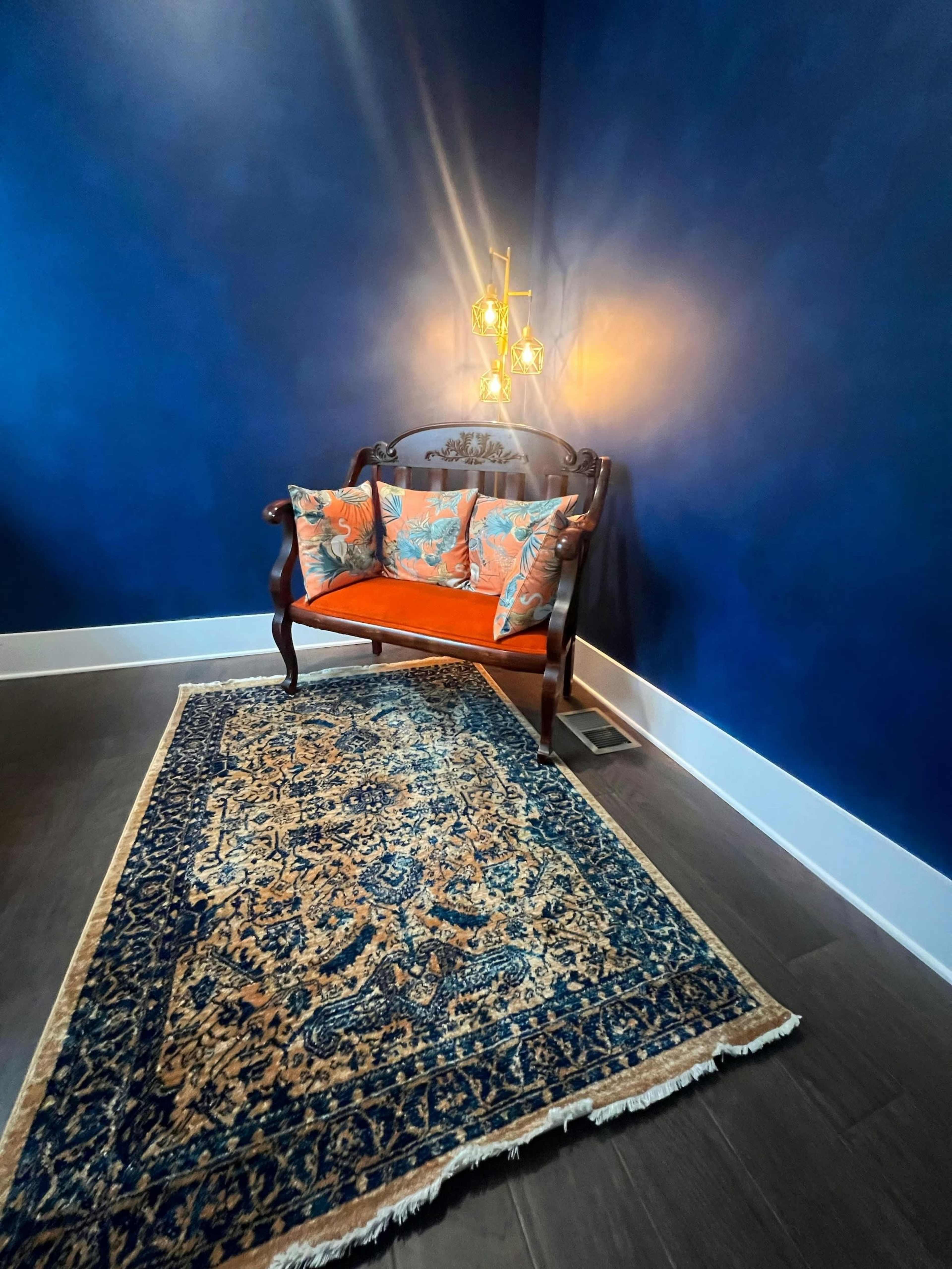 A wooden loveseat with orange cushions is positioned on a patterned rug against a dark blue wall, illuminated by a hanging light fixture.