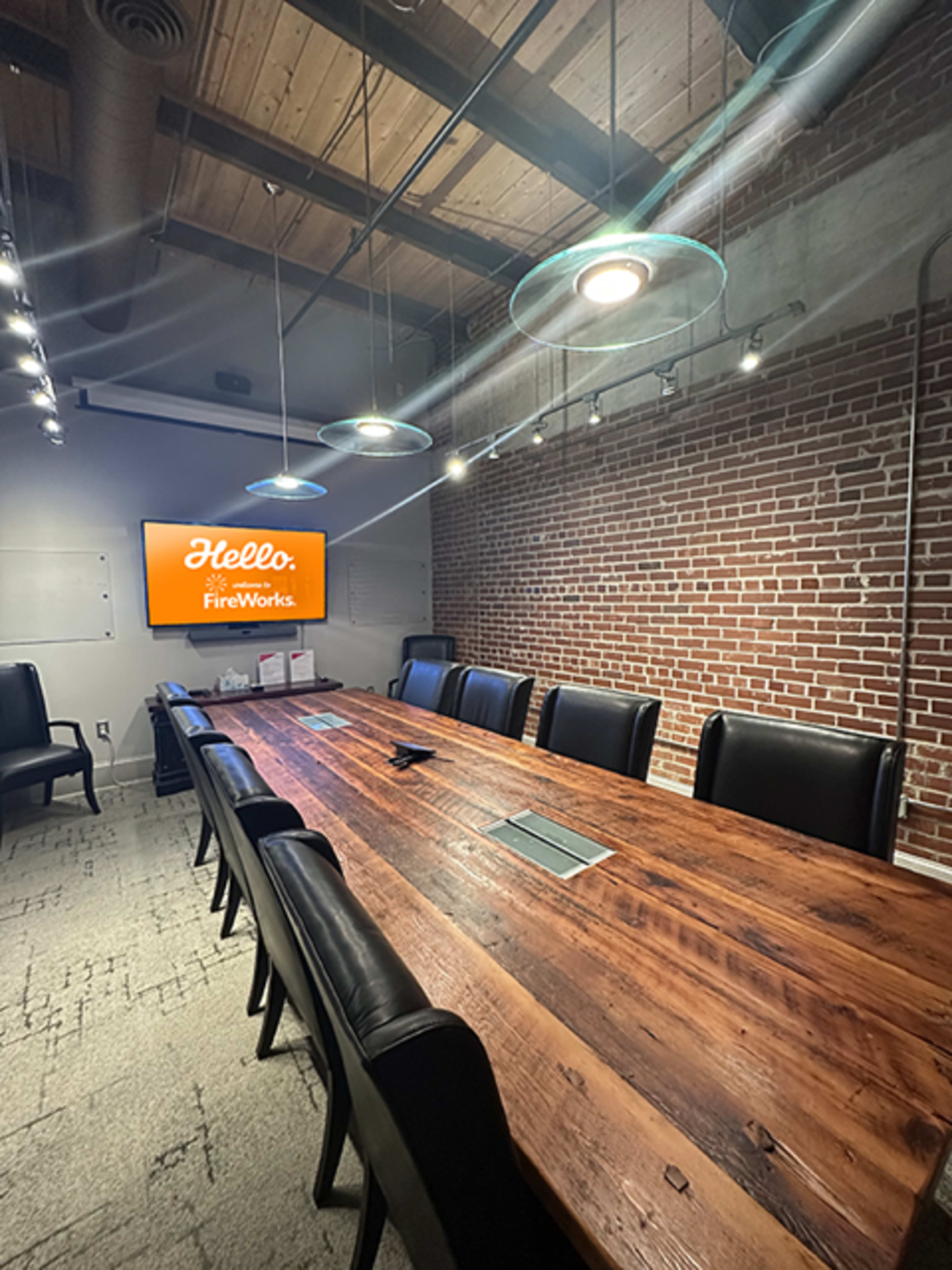 A long wooden conference table with black chairs is set up in a well-lit meeting room featuring exposed brick walls and a screen displaying "Hello" and "FireWorks."