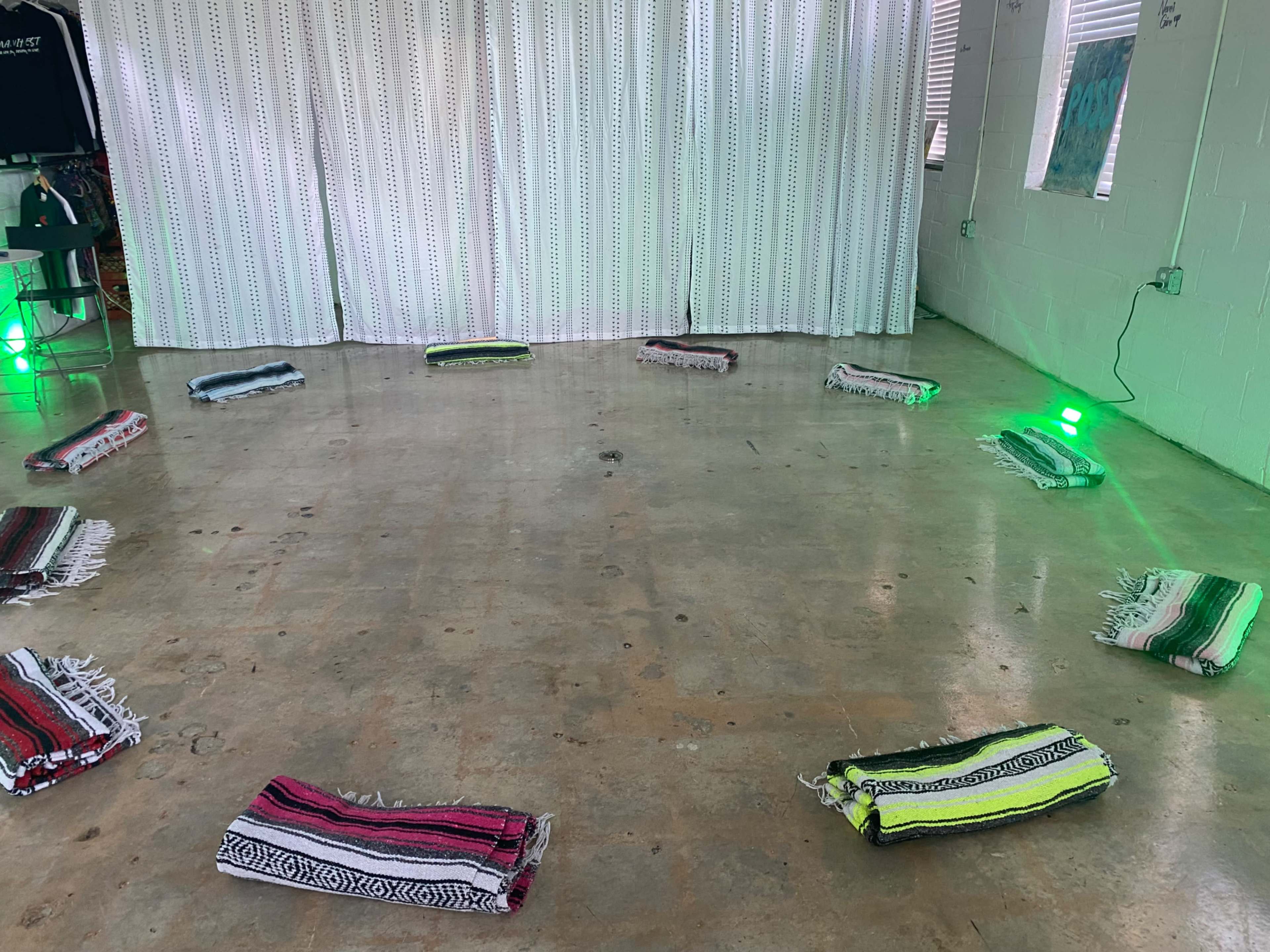 The image shows a circular arrangement of colorful blankets on a concrete floor, surrounded by green lights and a white patterned curtain.