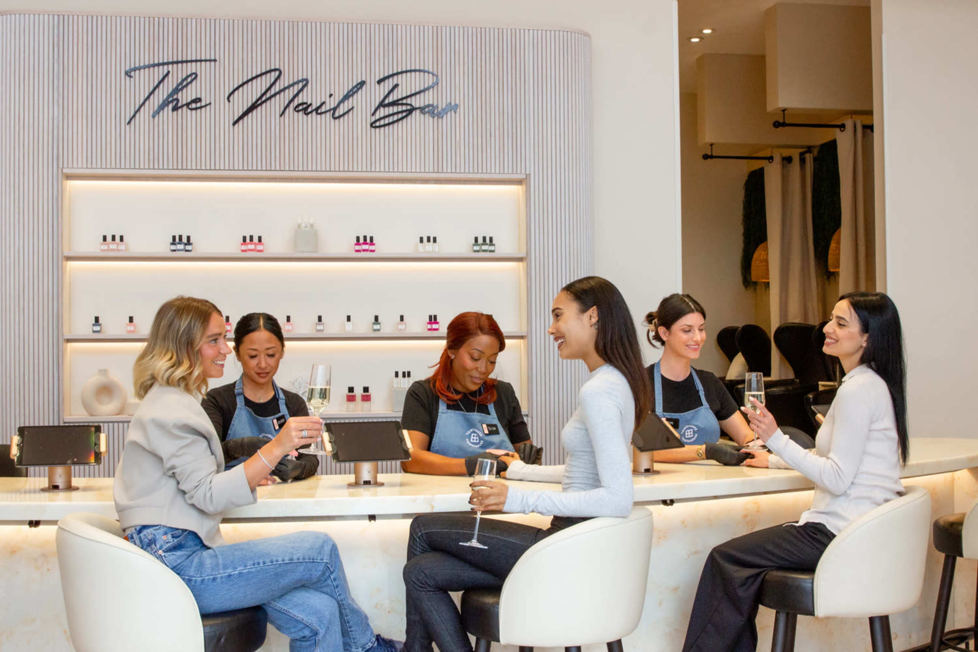 A group of women enjoy drinks while seated at a bar in a nail salon, with employees providing services in the background.