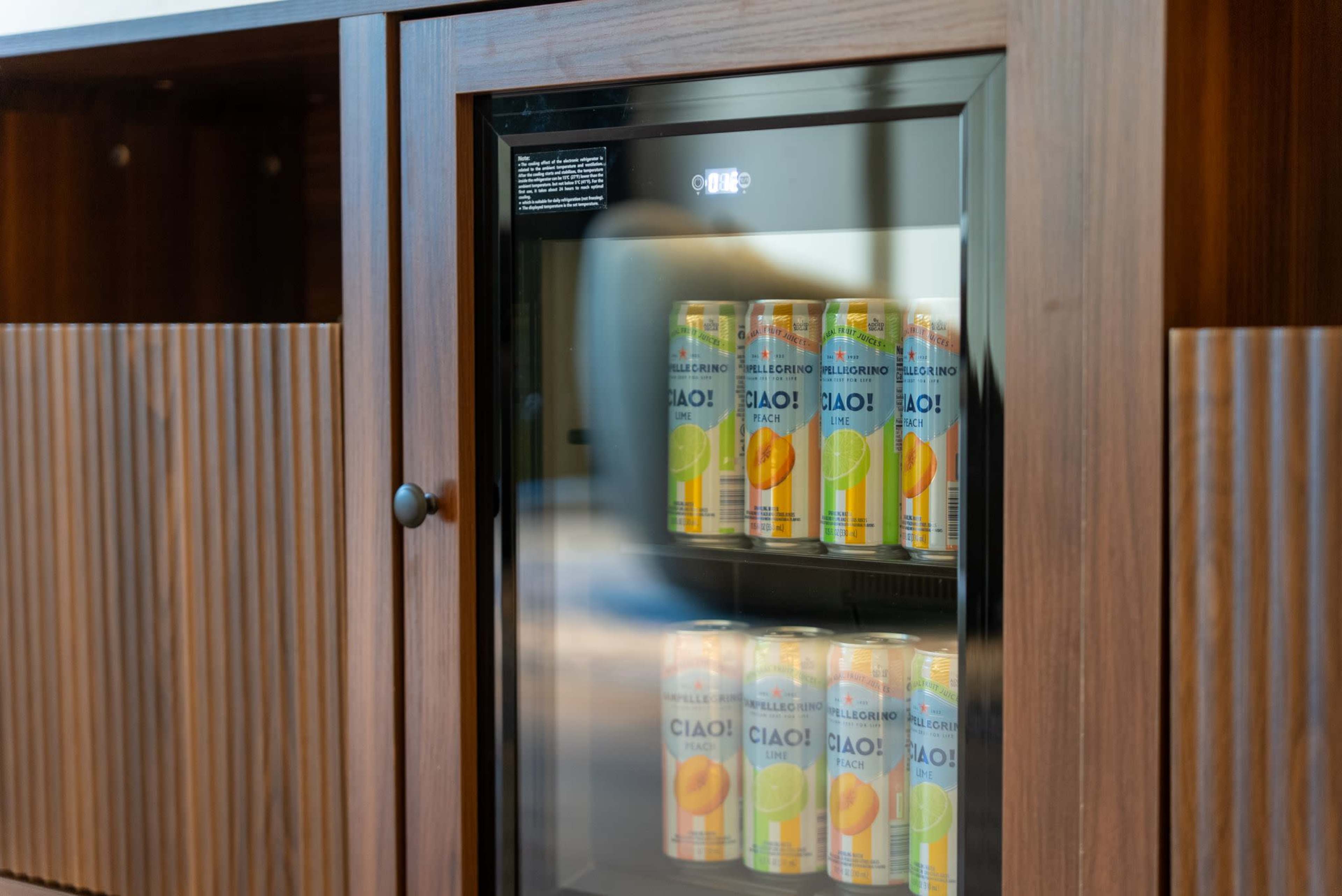The image shows a glass-fronted beverage refrigerator containing several cans of "Ciao!" fruit-flavored drinks, placed within a wooden cabinet.