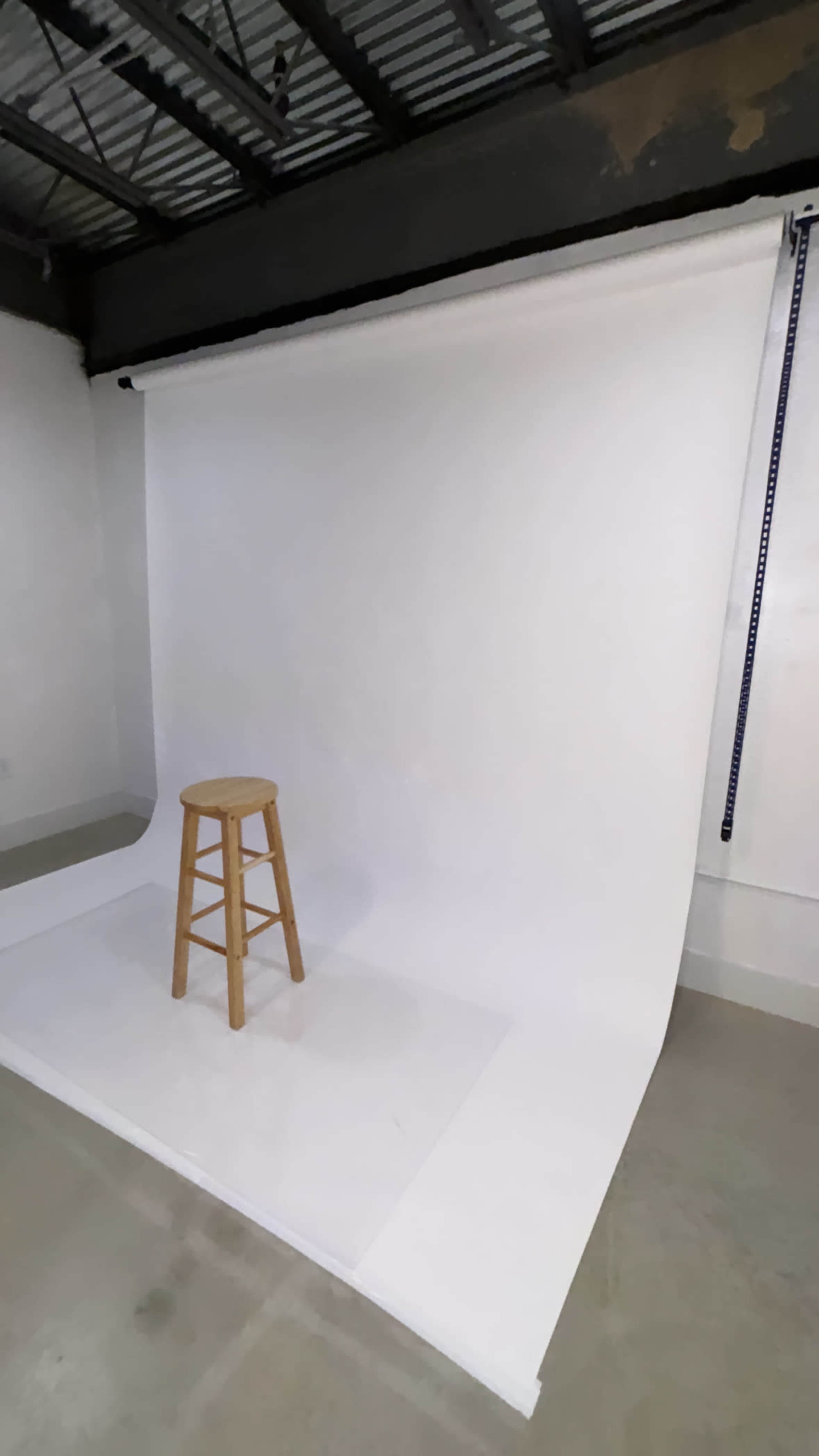 A wooden stool sits on a white backdrop in a studio space with gray flooring.
